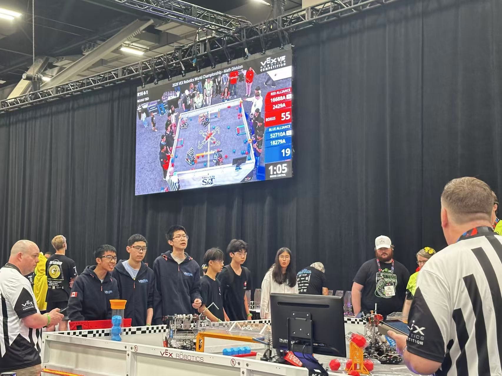 A row of clubbers standing along the barrier at a competition, watching a VEX IQ match on the screen above the field.