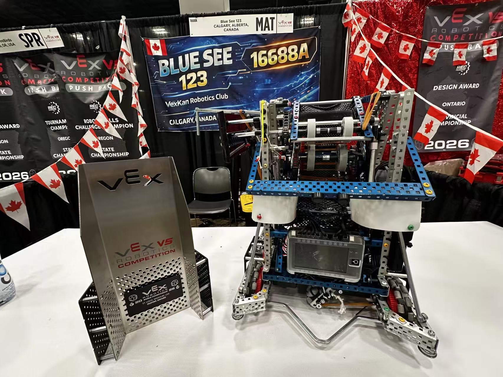 A metal V5 robot on the pit table beside the team's VEX award, Canadian flags strung across the booth behind it.