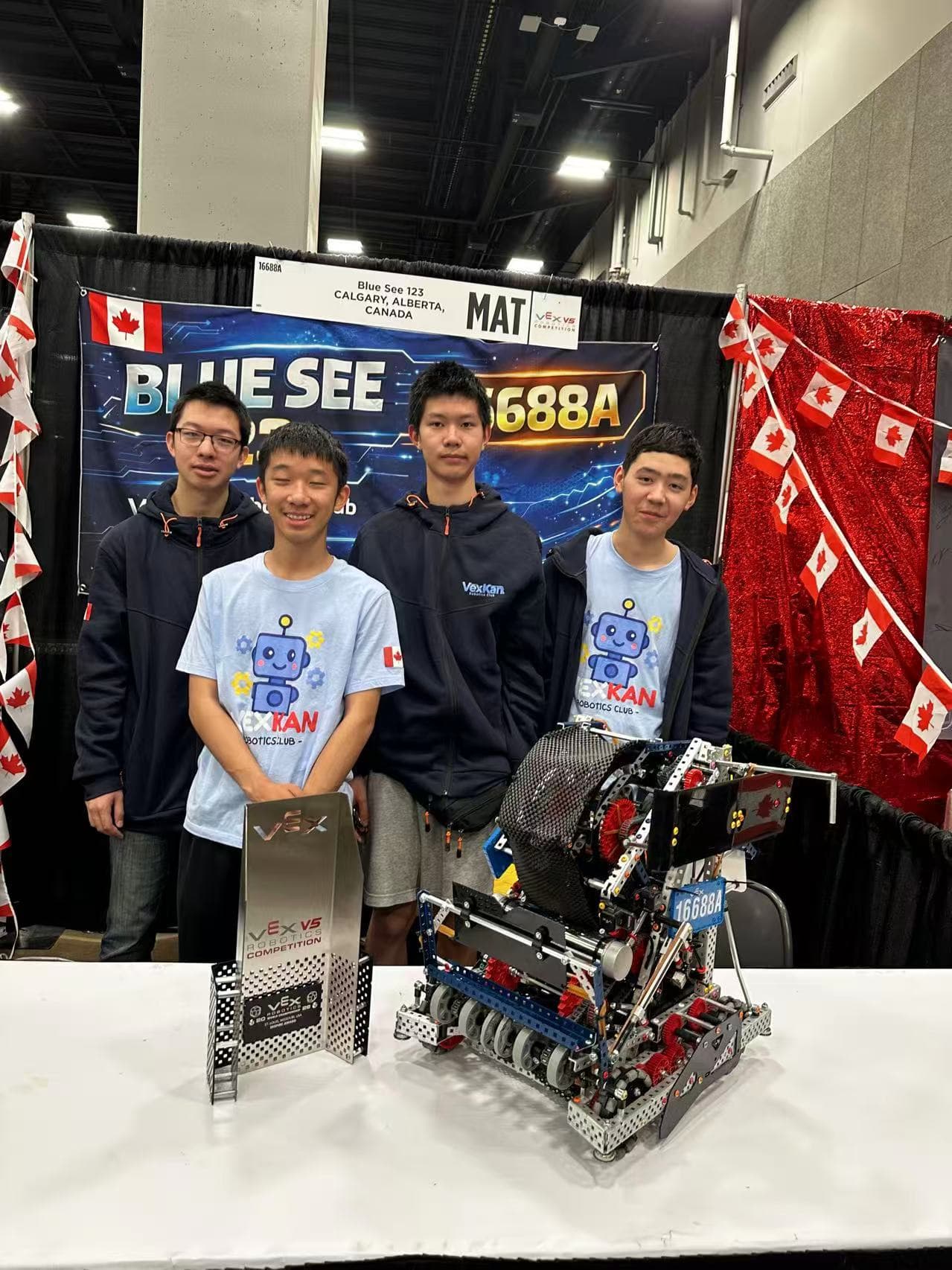Four clubbers in club shirts standing behind their V5 robot at their pit, under the team's banner.