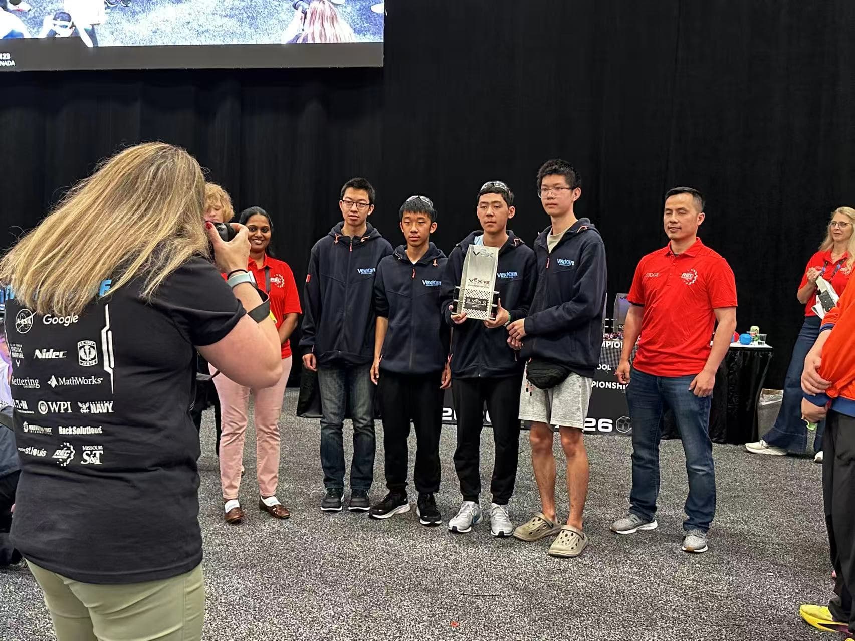 A group of clubbers standing together on the competition floor while someone photographs them, one holding the team's robot.