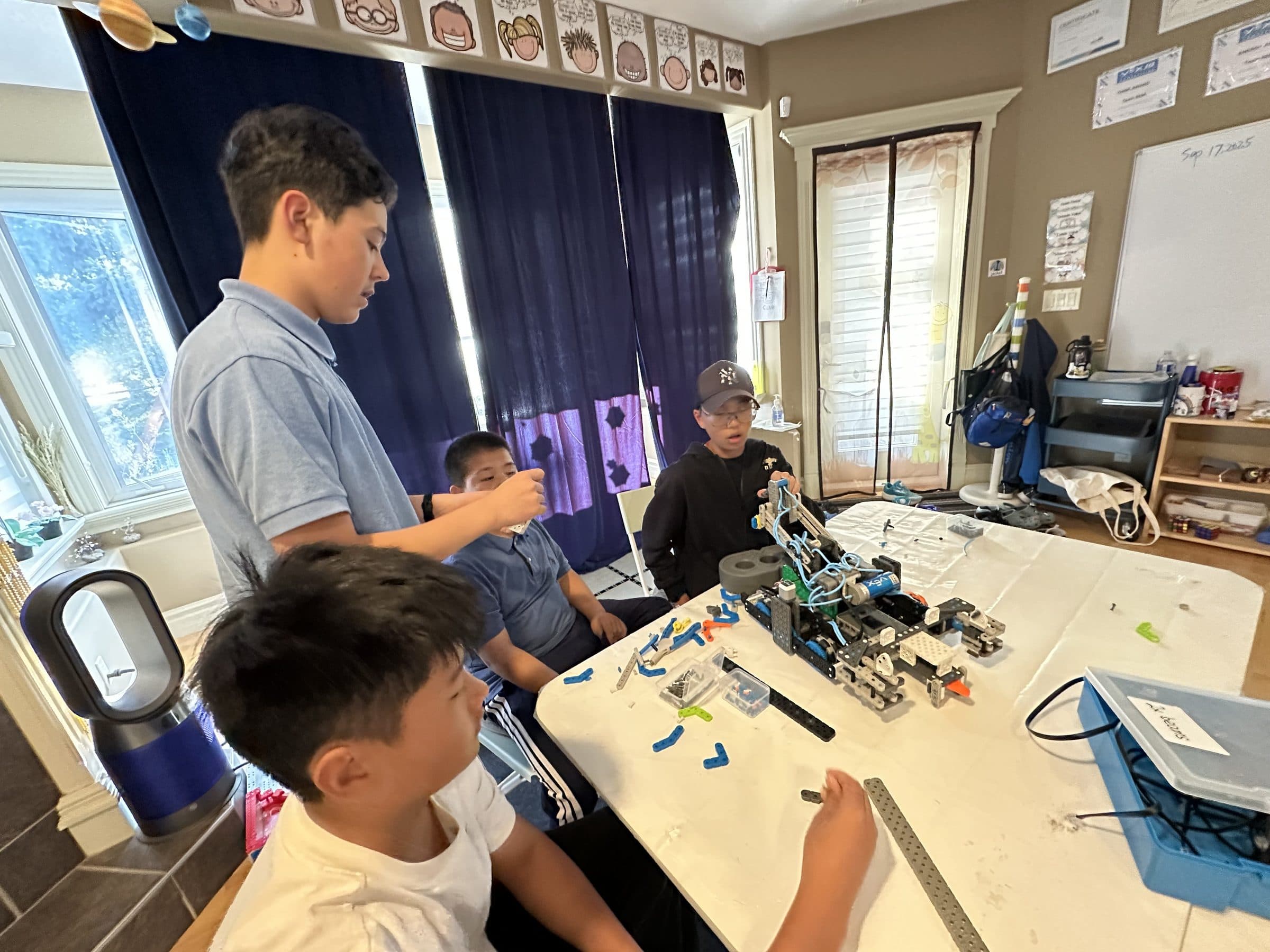 Four clubbers around a table at a build session, a part-built VEX IQ robot and sorted parts in front of them and the session's date written on the whiteboard behind.