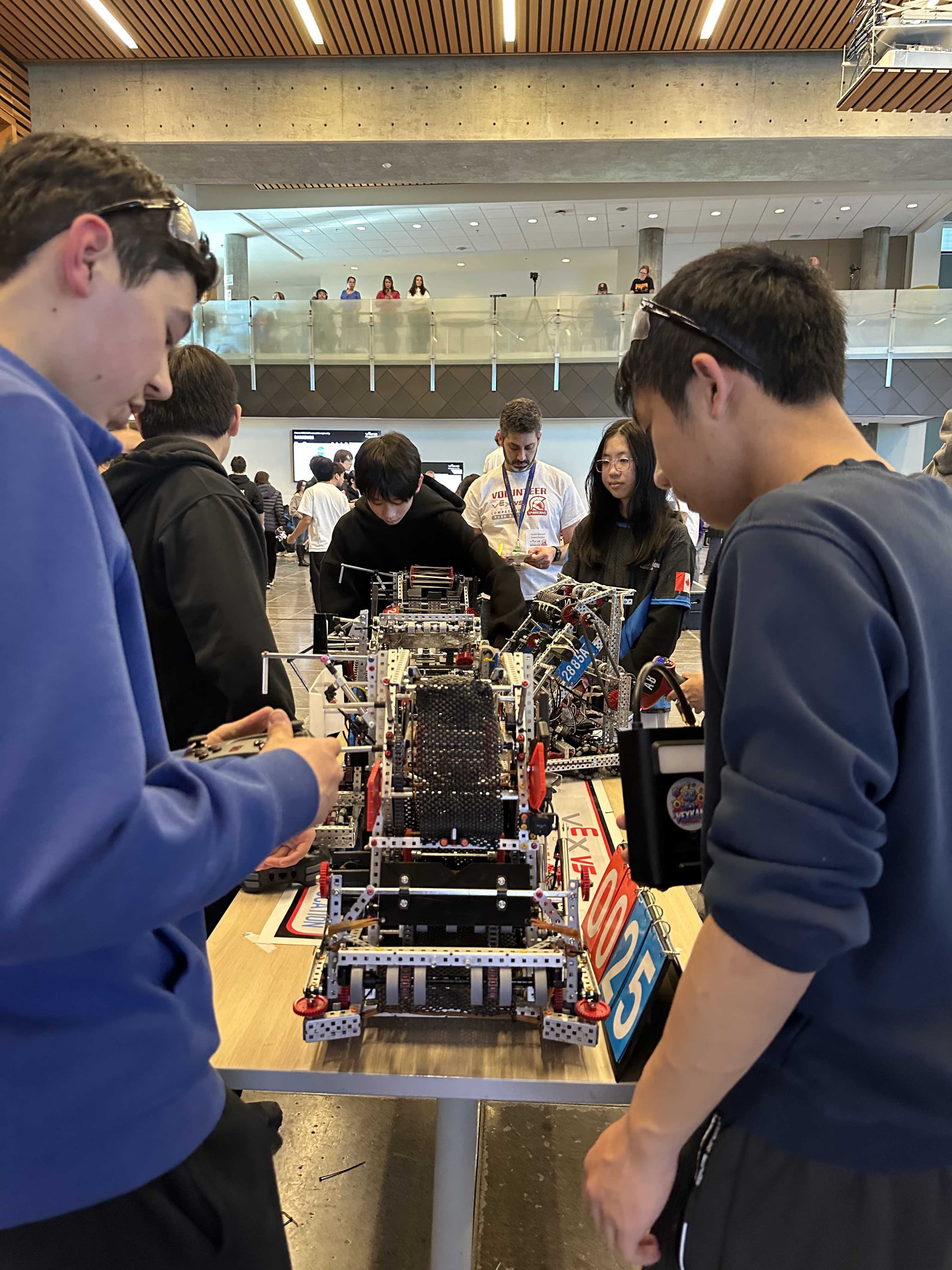 Two students lifting a V5 robot onto a table in the bright atrium of a competition venue.