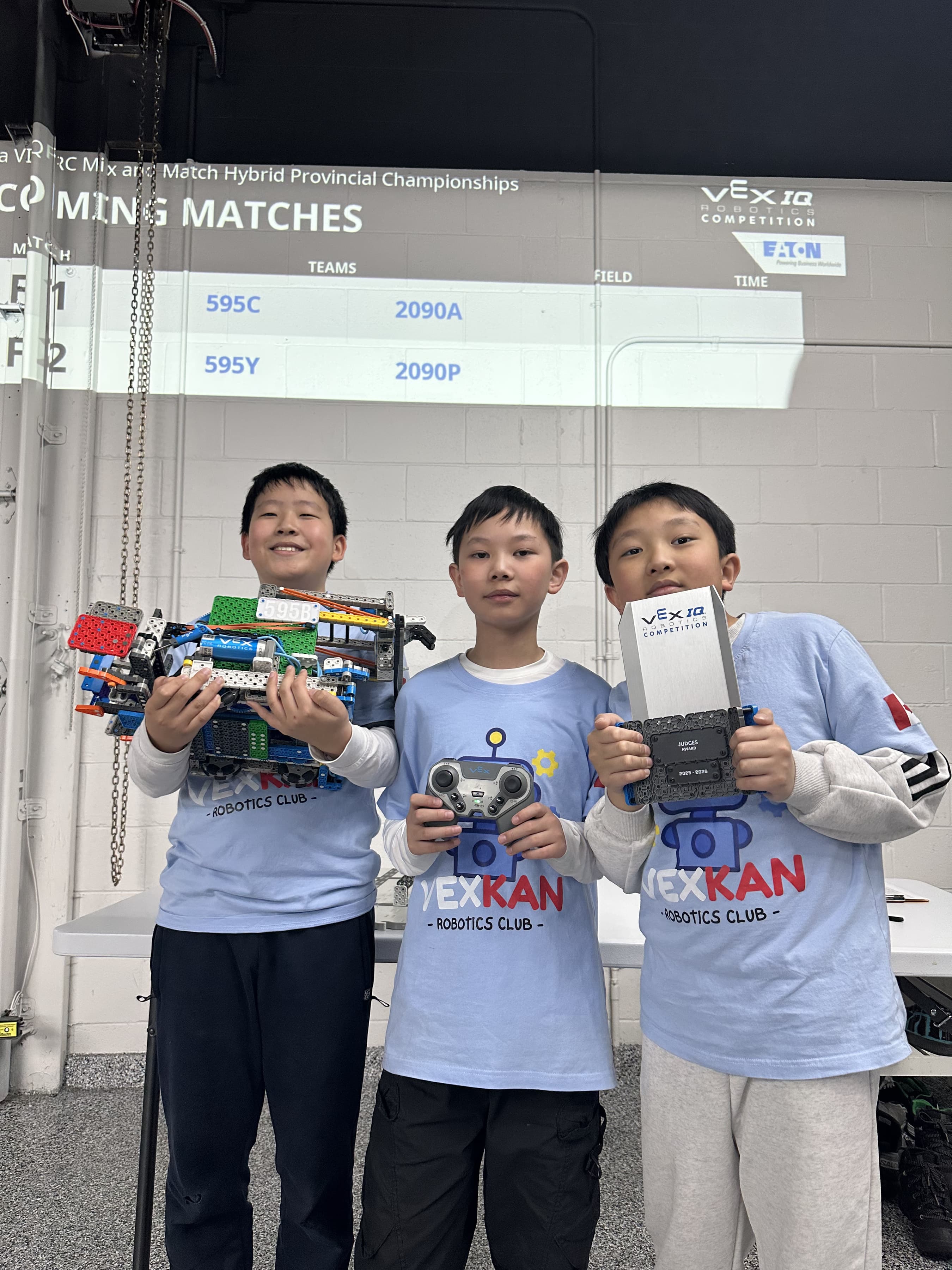 Three members of 595B in club shirts, one holding their VEX IQ robot, one a controller and one the Judges Award trophy.