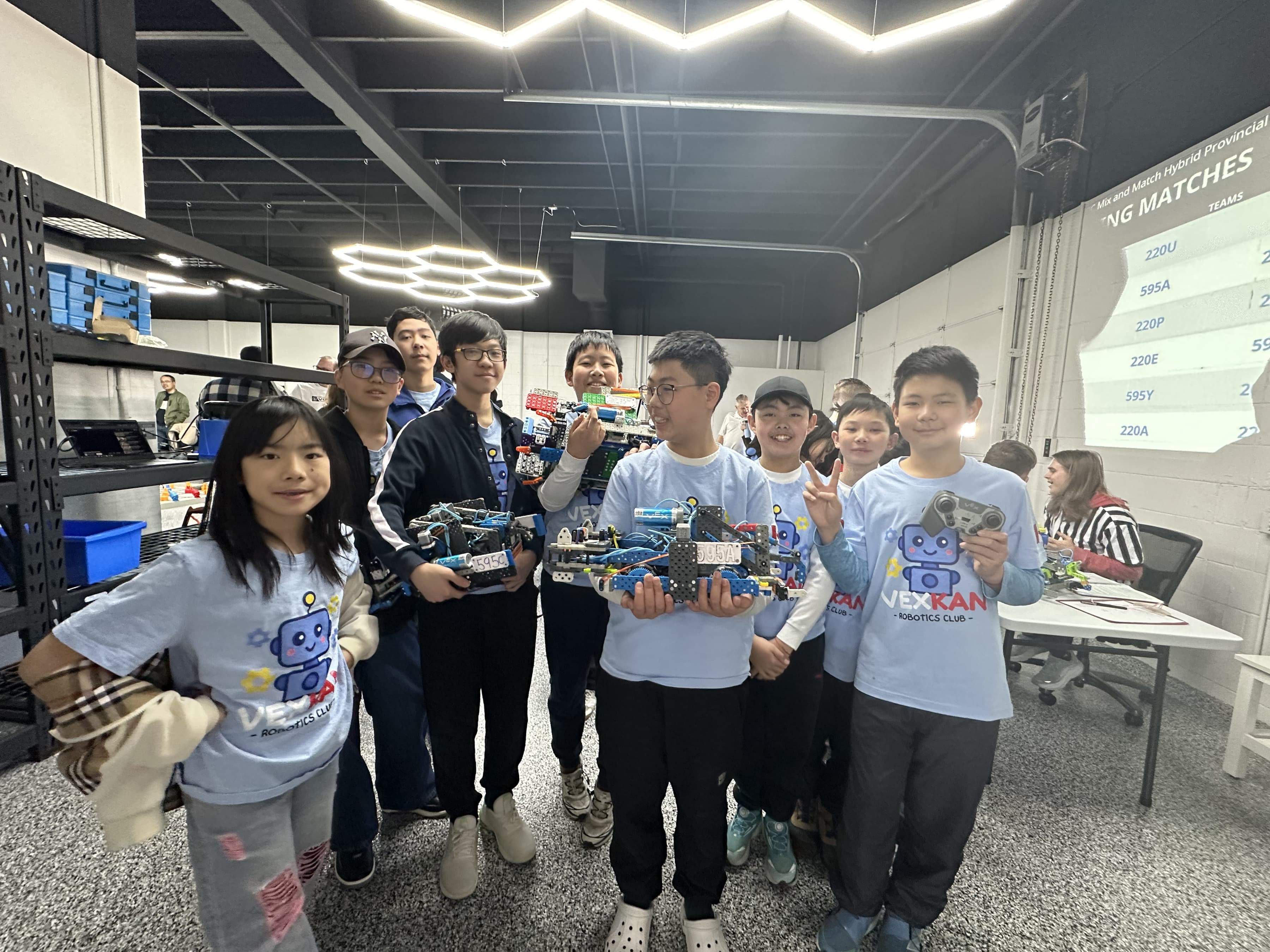 Nine clubbers in club shirts holding their VEX IQ robots and controllers at the provincial championships, the match schedule projected on the wall behind them.