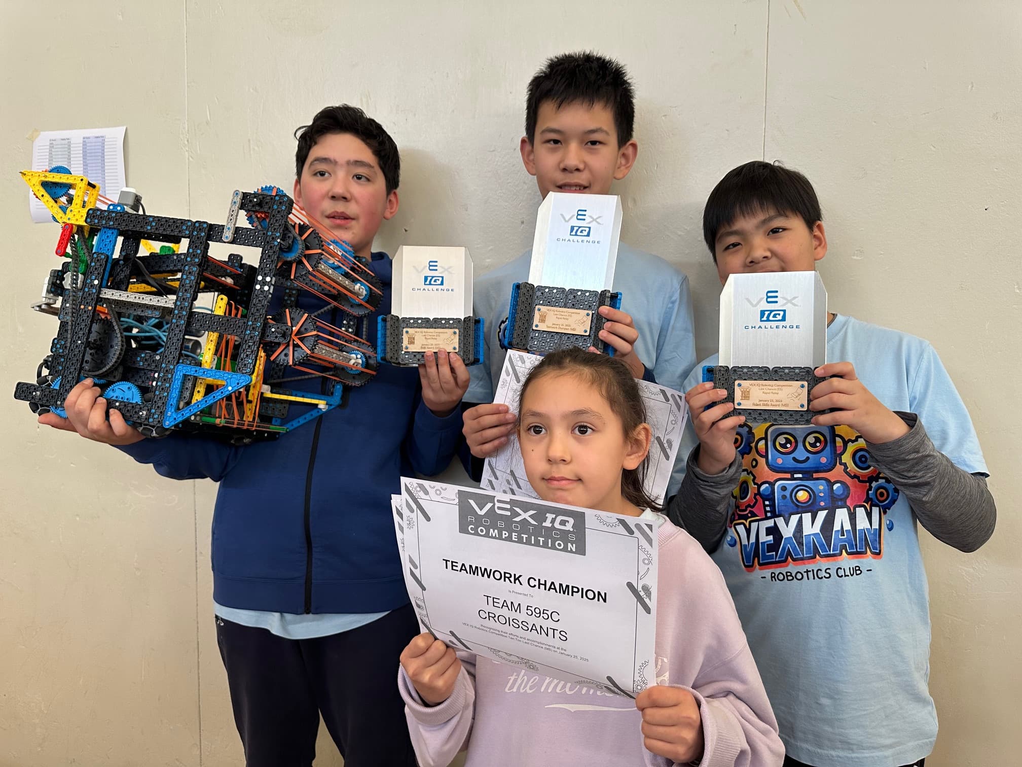 Three members of 595C with their VEX IQ trophies and a Teamwork Champion certificate lined up on the table in front of them.