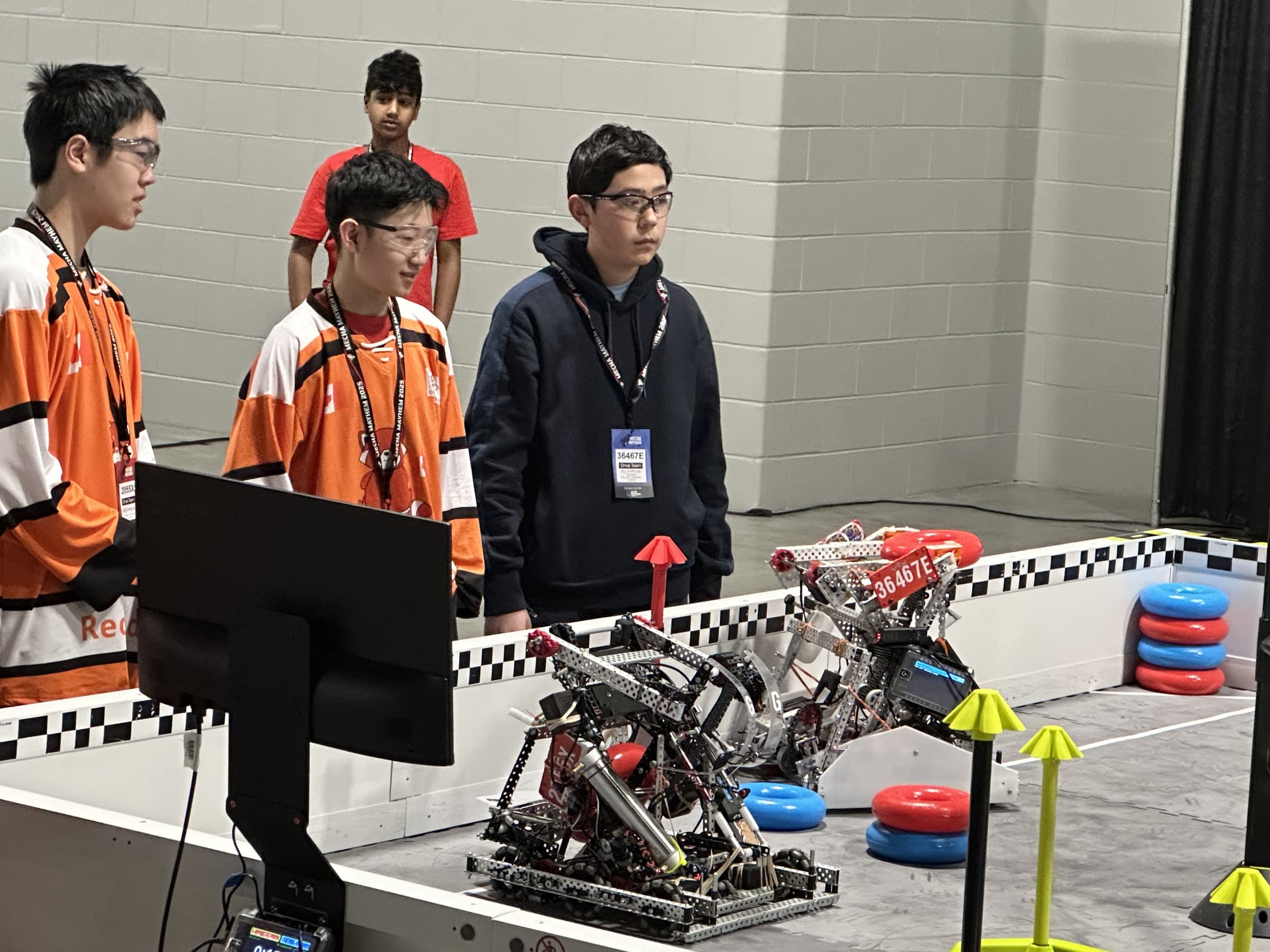 Three drive team members standing at the field wall in team jerseys, watching 36467E's robot and an opposing robot on the tiles below.