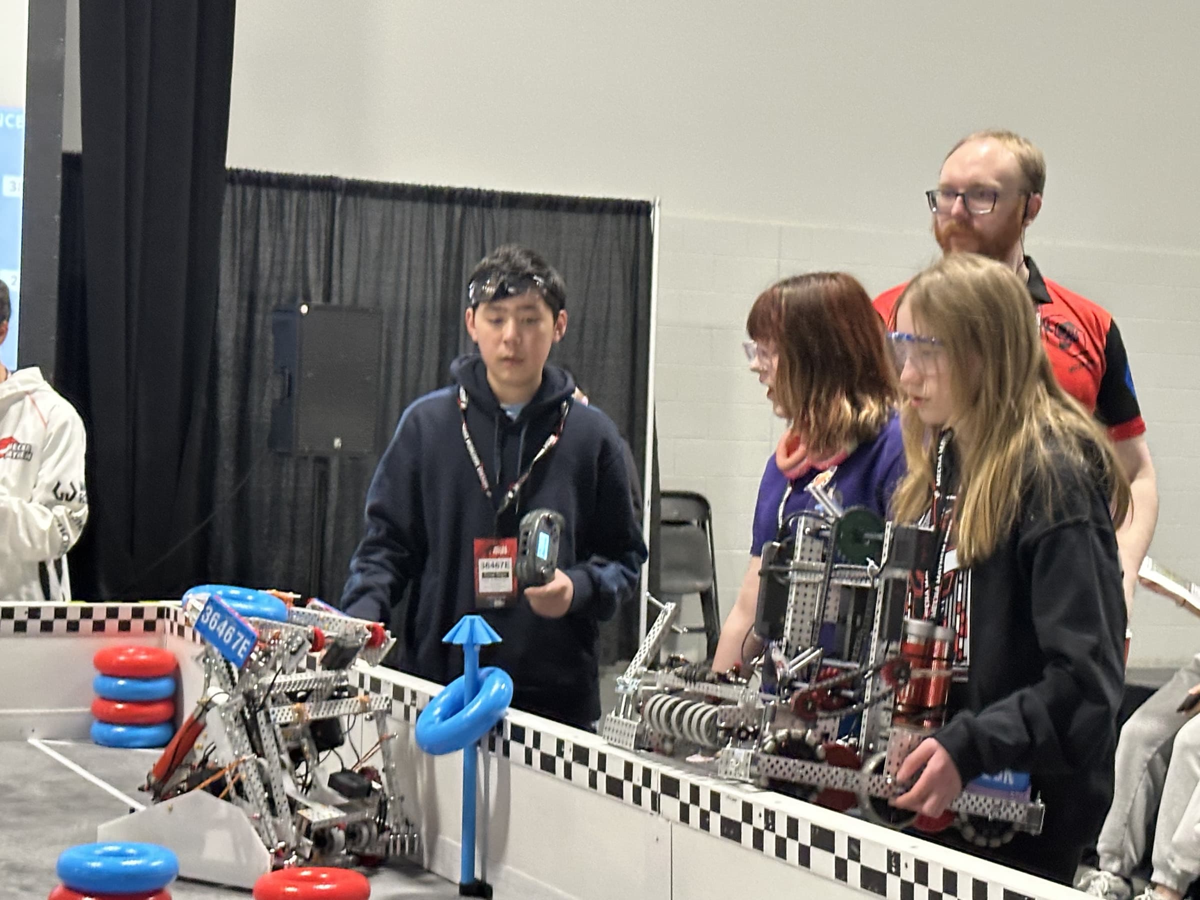 Drivers from three teams lined up along the barrier of a V5 field, 36467E's robot on the tiles in front of them and their alliance partners' robots being set down alongside.