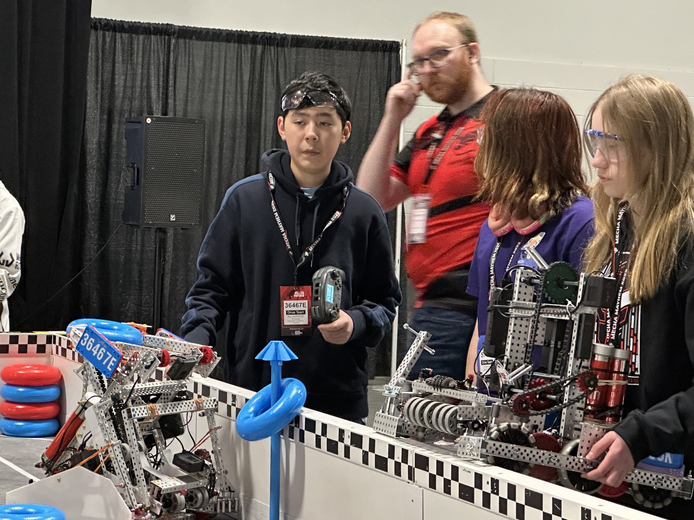 A 36467E driver holding a controller at the edge of the field, drive teams from two other clubs lined up along the wall beside him.