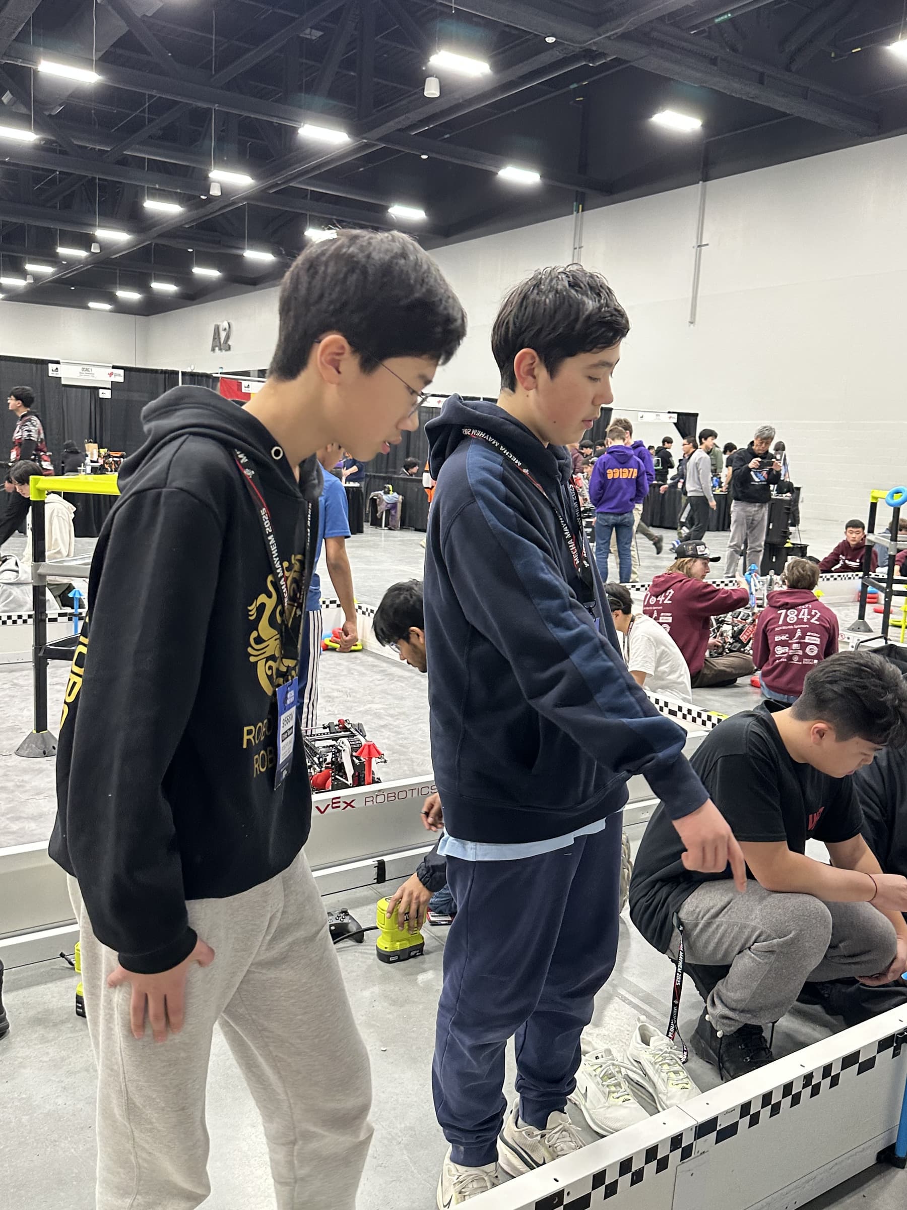 Two drive team members standing on the field wall looking down at the tiles, the rest of the competition hall and other clubs' pits stretching out behind.