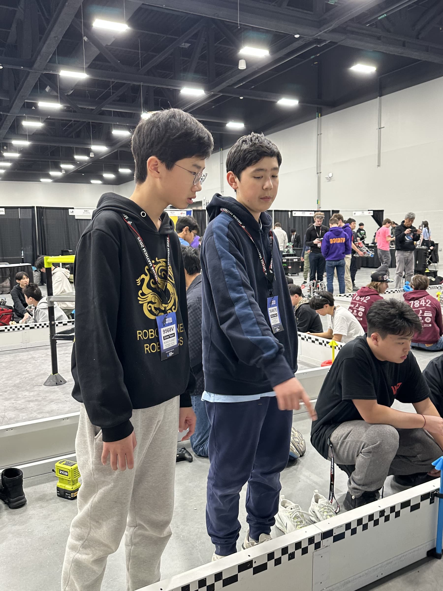 Two drive team members standing up on the field barrier at Mecha Mayhem, looking down at the field while other teams work at the pits behind them.