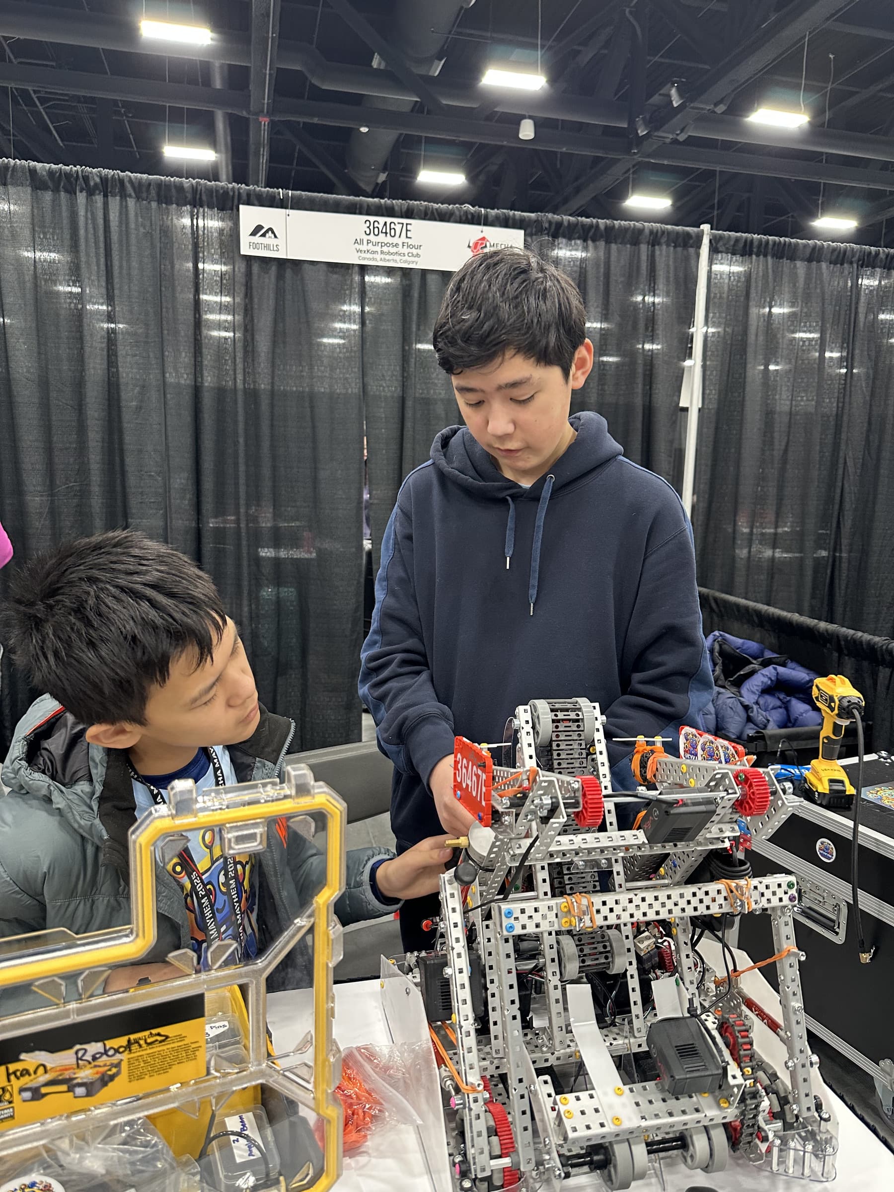 A clubber fitting the 36467E number plate onto the team's metal V5 robot at the pit table, while another watches from across the bench.