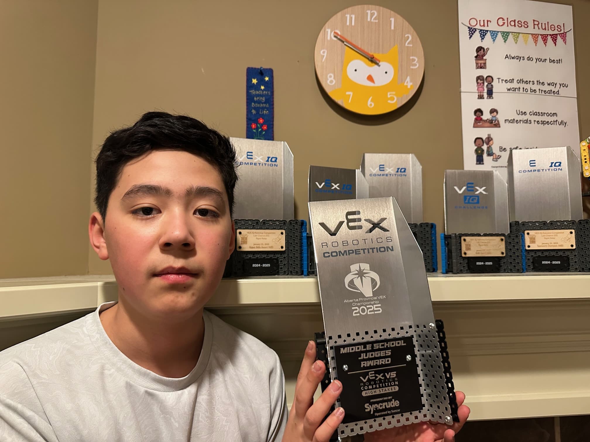 A member of 36467E holding the Middle School Judges Award from the Alberta V5 provincial championship, the club's other trophies on the shelf behind.