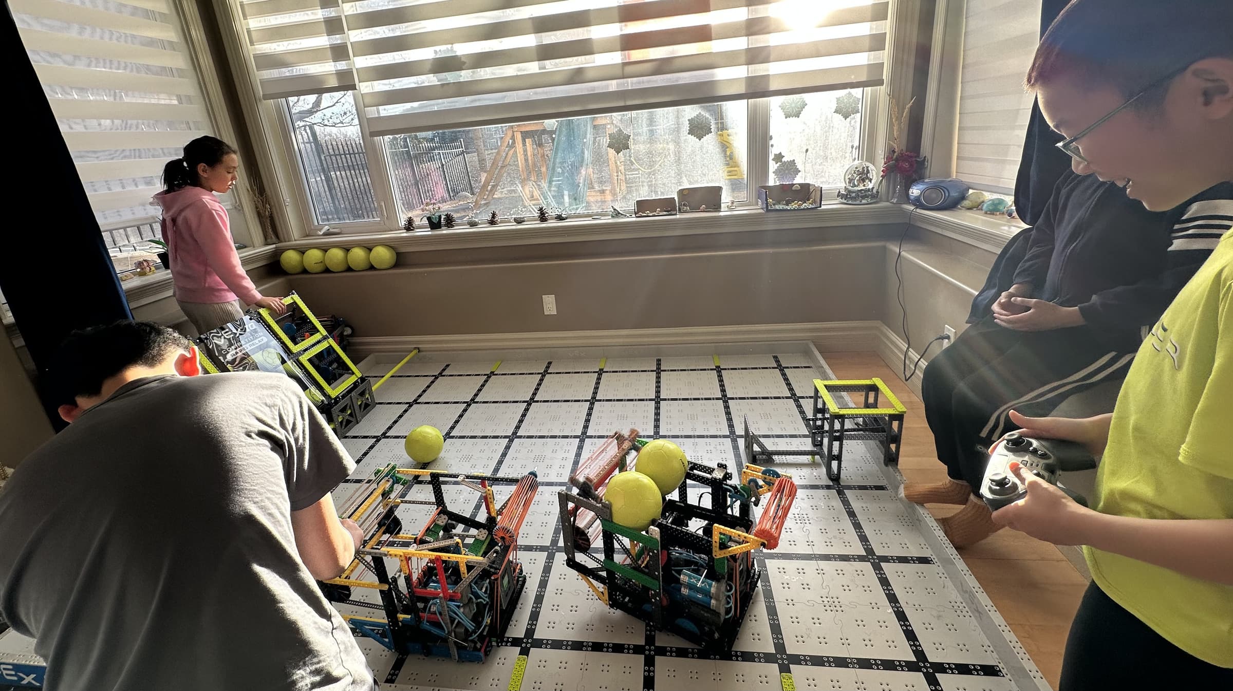 Four clubbers around a VEX field laid out on the floor of a front room, two robots and several game balls on the tiles, one of them driving.