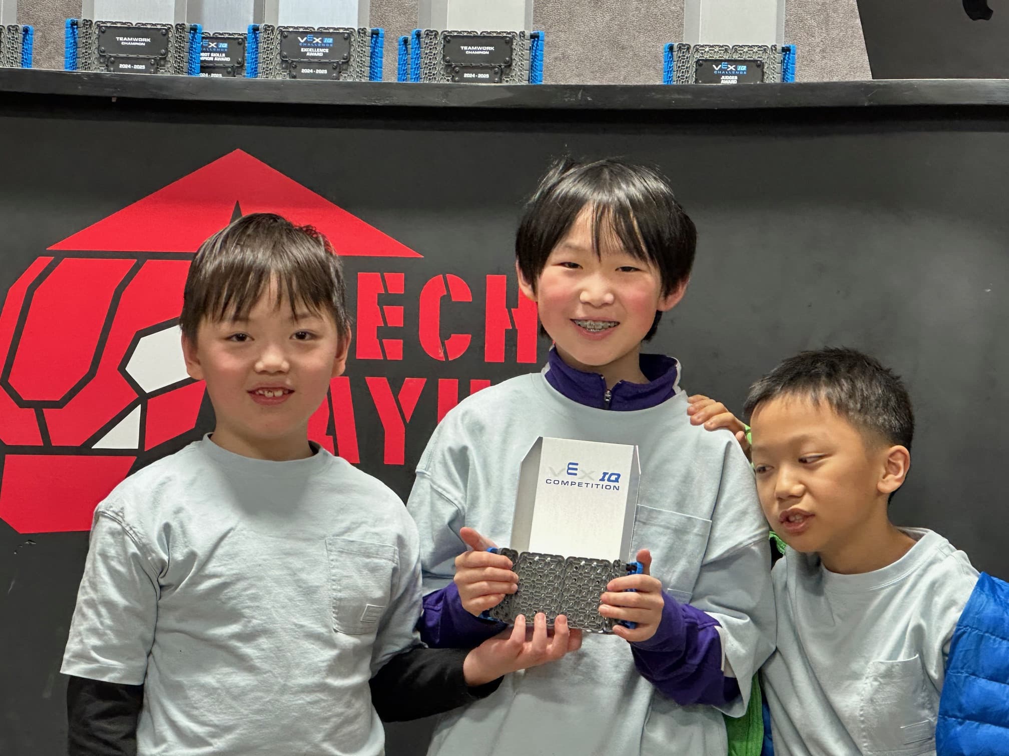 Three young clubbers holding a VEX IQ Competition trophy in front of the Mecha Mayhem backdrop, a shelf of the event's other trophies above them.