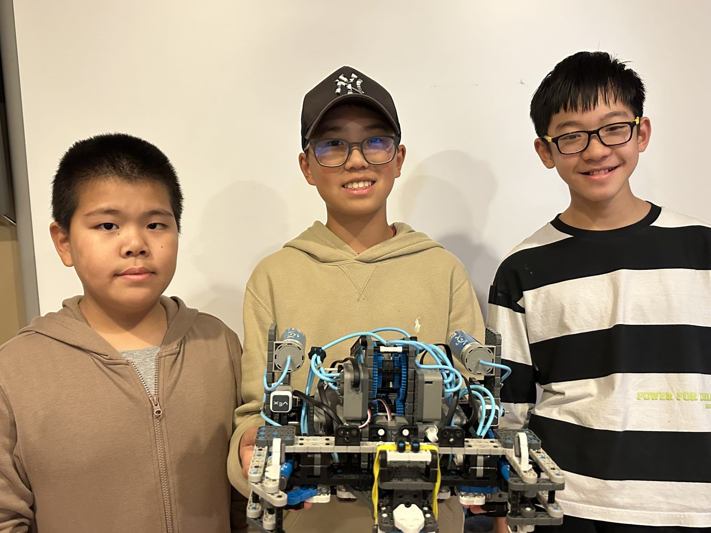 Three clubbers holding up the VEX IQ robot they have just finished, its motors and cabling visible along the front of the chassis.