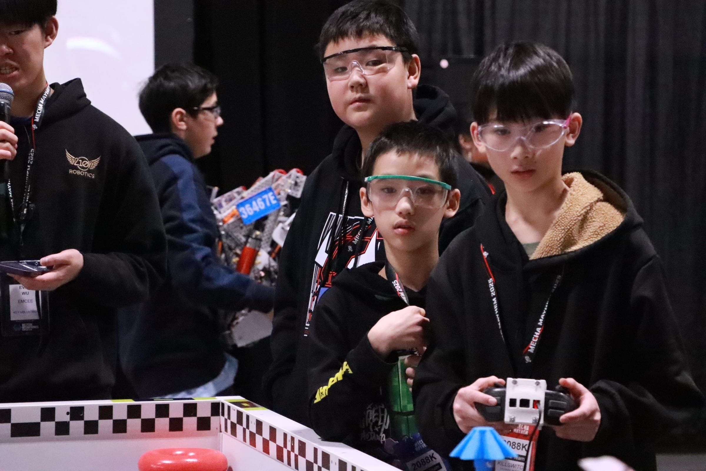 Three young drivers in safety glasses at the field barrier, one holding a controller, with 36467E's robot on the table behind them and an emcee with a microphone to one side.