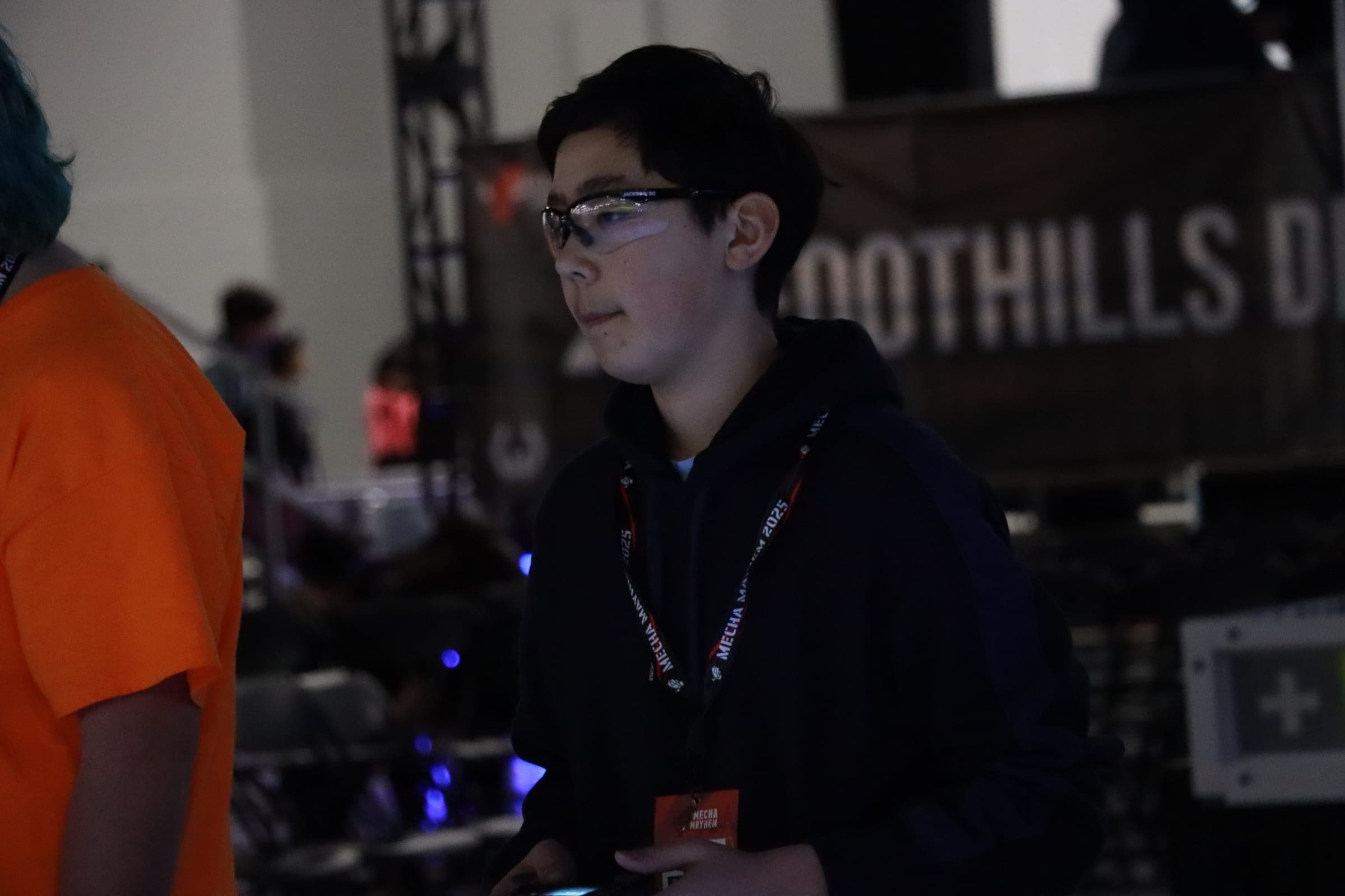 A driver in safety glasses and a competition lanyard watching the field intently, controller held low, the hall dark behind him.