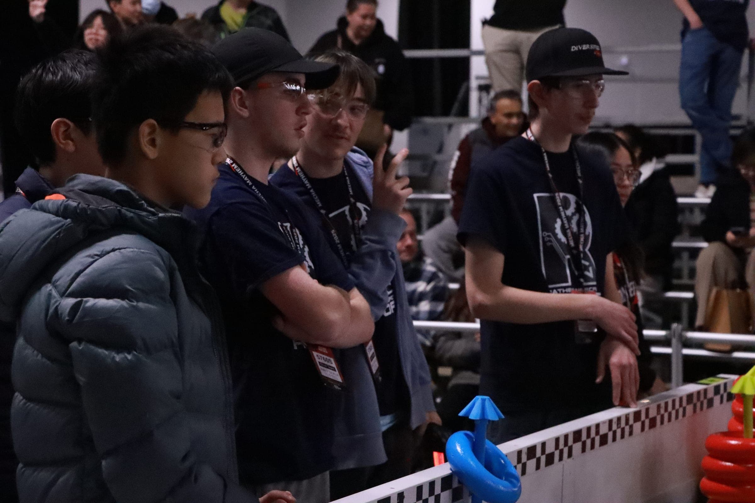 Drivers from several teams standing shoulder to shoulder along the field barrier in a darkened hall, watching the match in front of them.