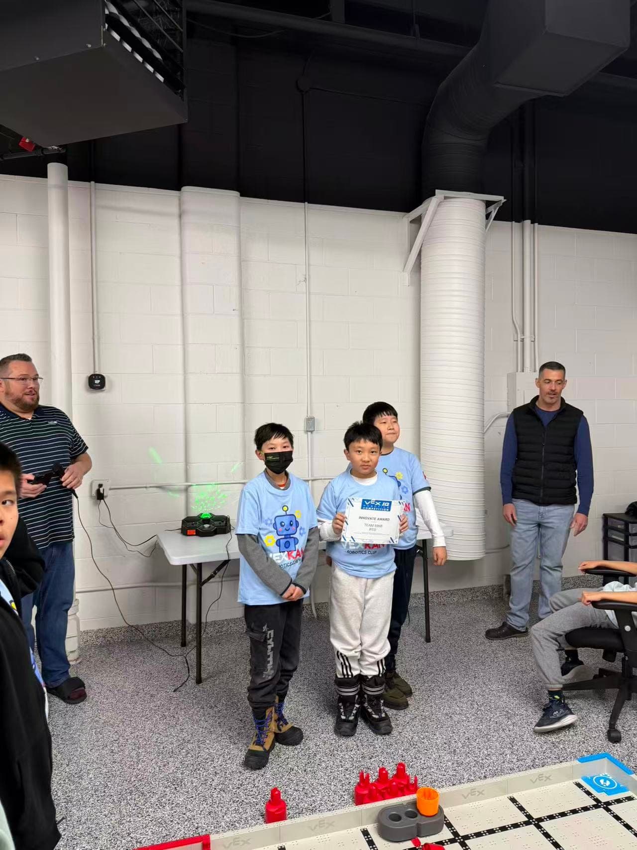 Three young members of 595B standing together in club shirts, the one in front holding their Innovate Award certificate, with the VEX IQ field in the foreground.