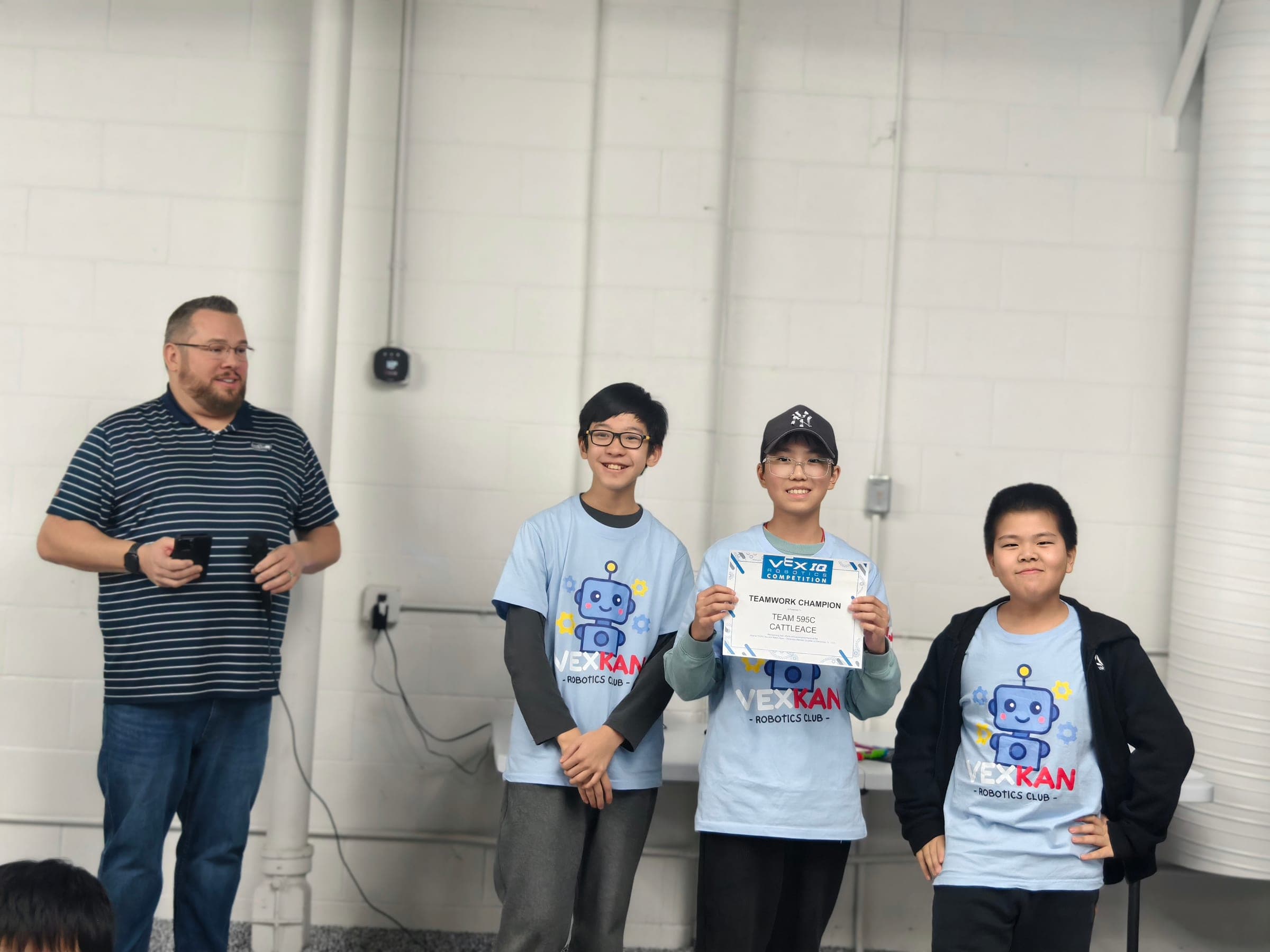 Three members of 595C in club shirts at the front of the room, the middle one holding up their Teamwork Champion certificate while a coach stands to one side.