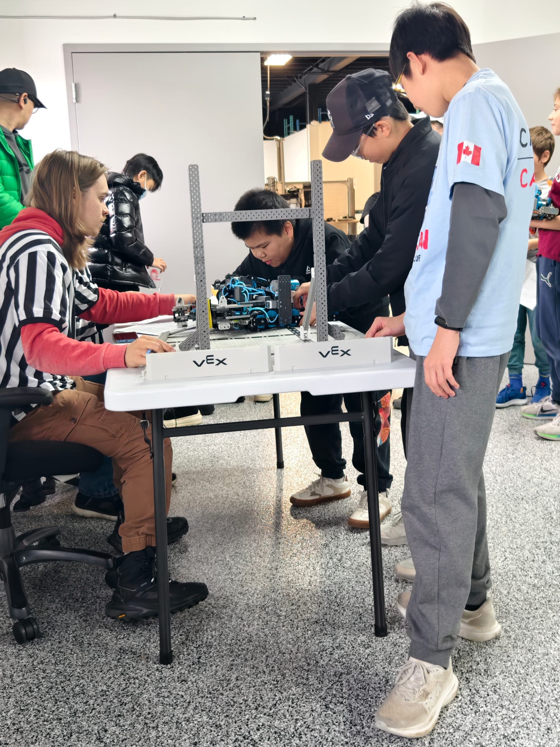 A referee seated at the inspection table measuring a V5 robot against the sizing tool while three students stand around the table waiting.