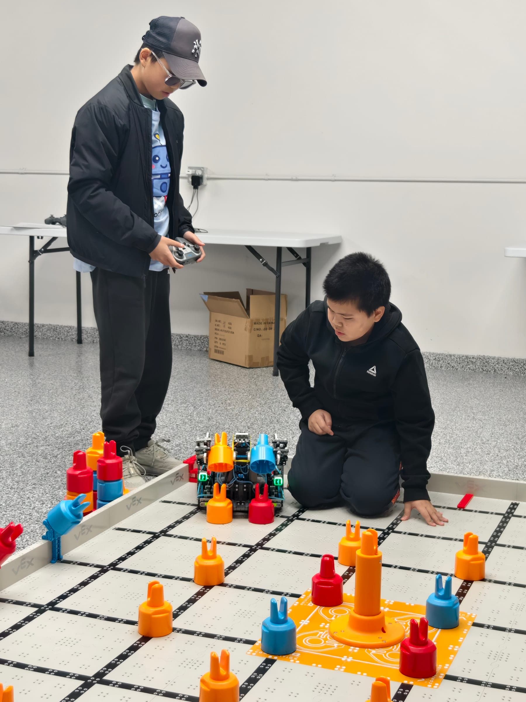 Two clubbers at a VEX IQ field laid out on the floor of the club room, one standing with the controller and the other kneeling at the edge of the tiles watching the robot.