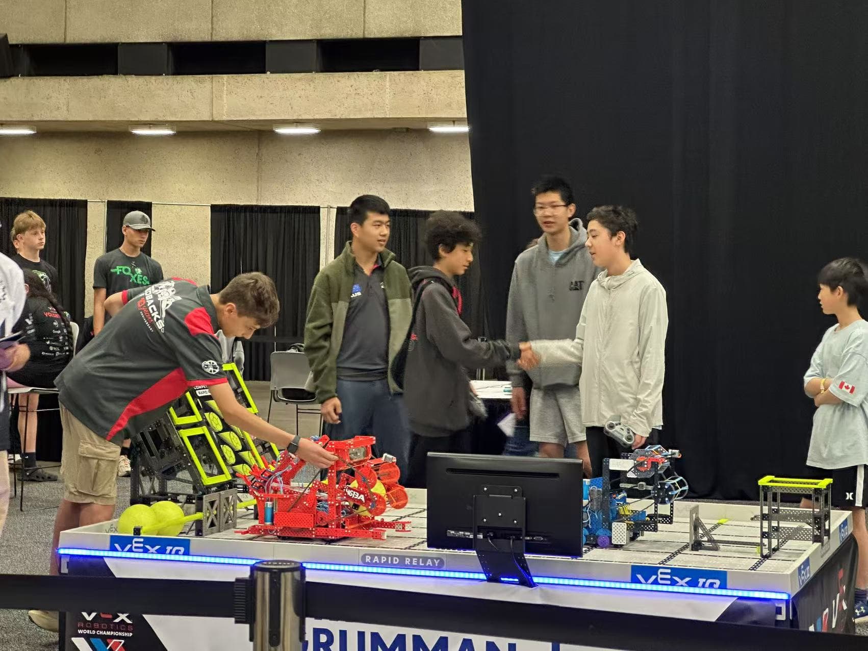 Two students shaking hands across a VEX IQ Rapid Relay field at the World Championship while another sets his robot down on the tiles.