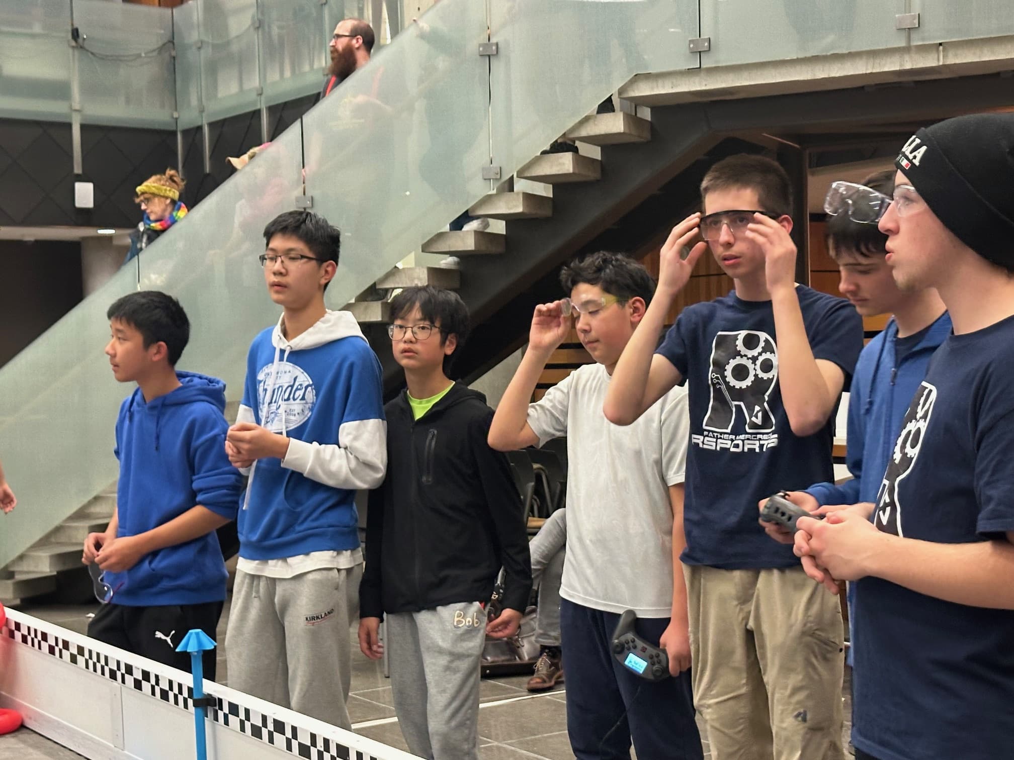 Clubbers lined up along the field barrier at a competition, one pulling on a pair of safety glasses.