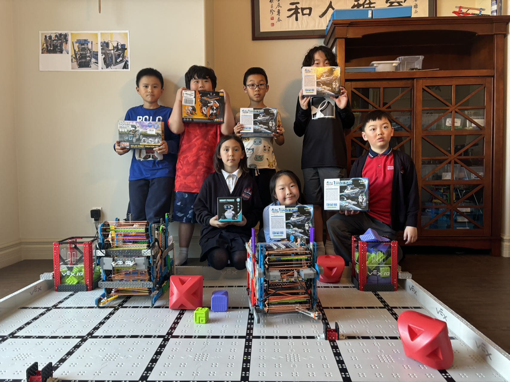 Young clubbers standing behind a VEX IQ field holding up their kit boxes and instruction books, their finished robots on the tiles.