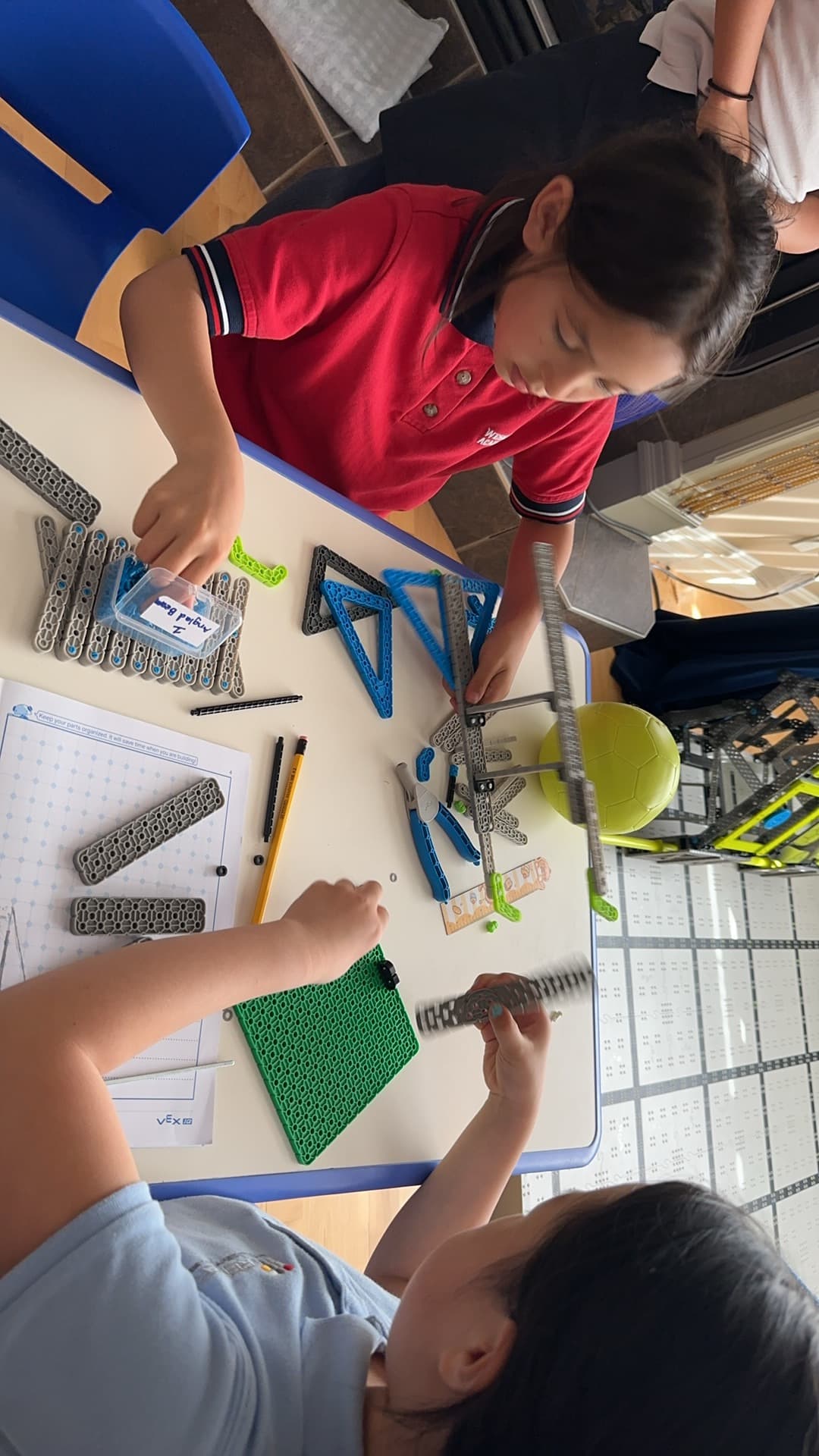 Two young clubbers building at a table, with VEX IQ beams sorted into rows and a parts sheet open beside them.
