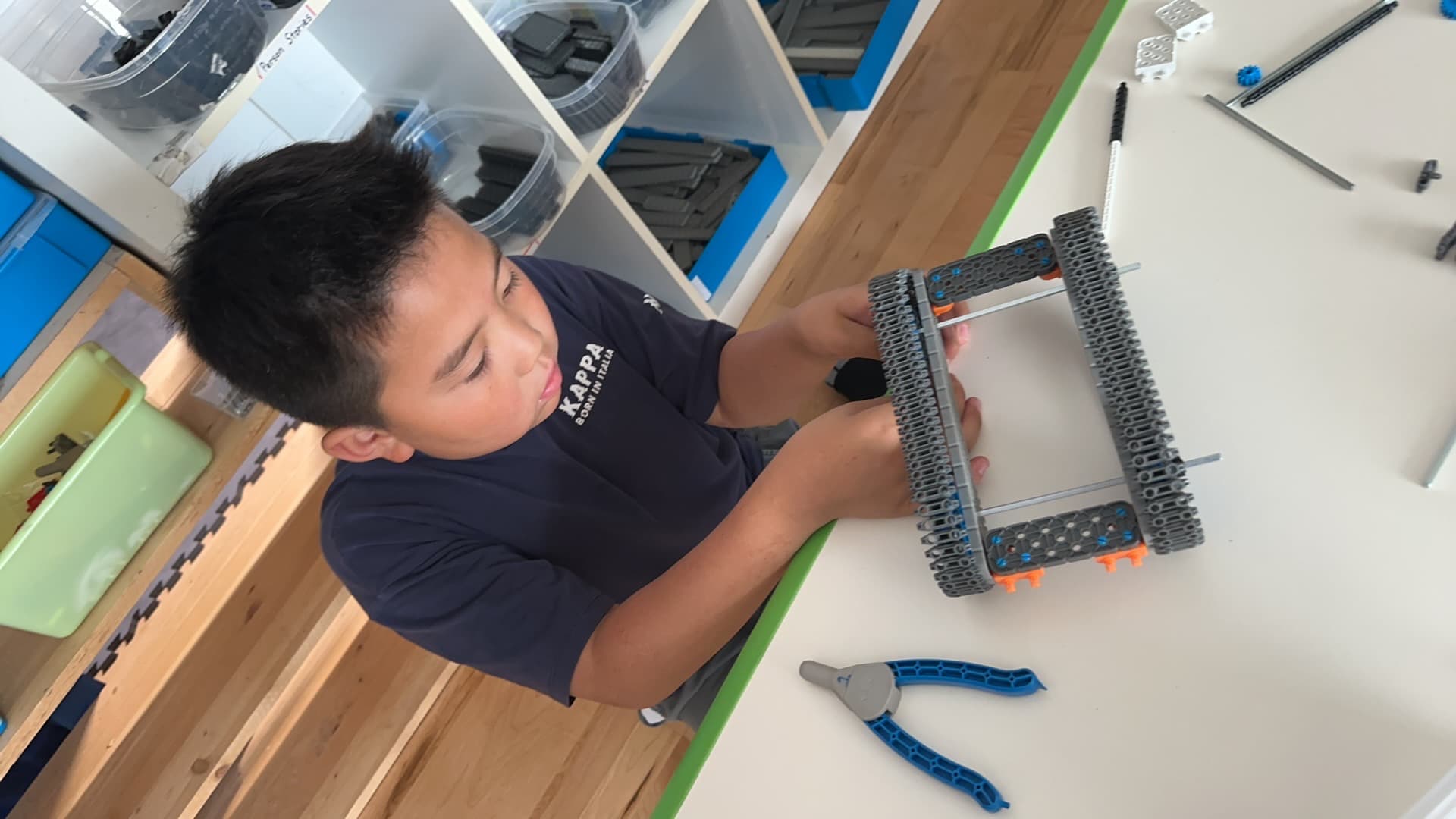 A clubber assembling a VEX IQ chassis at a workbench, a pair of pliers on the table beside him.