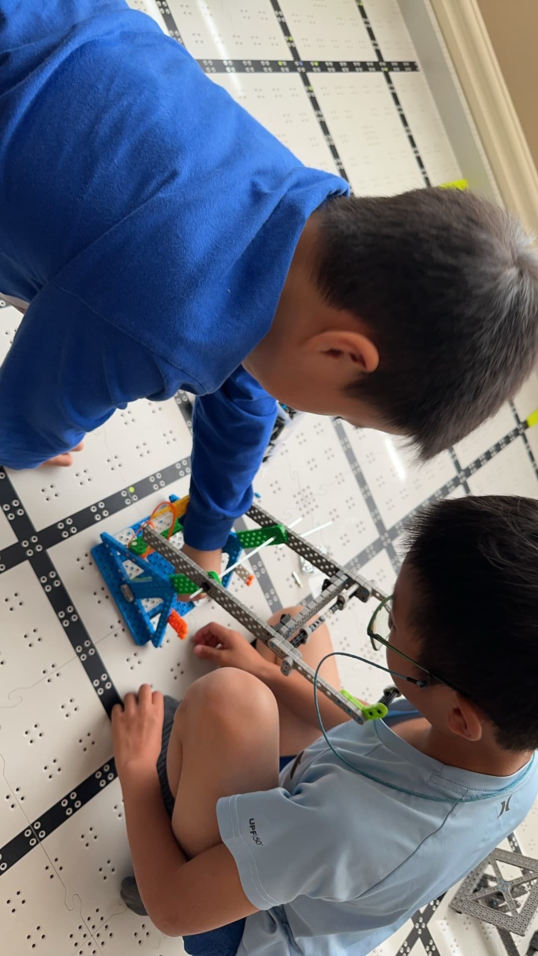 Two clubbers kneeling on a VEX IQ field mat, building a robot on the tiles.