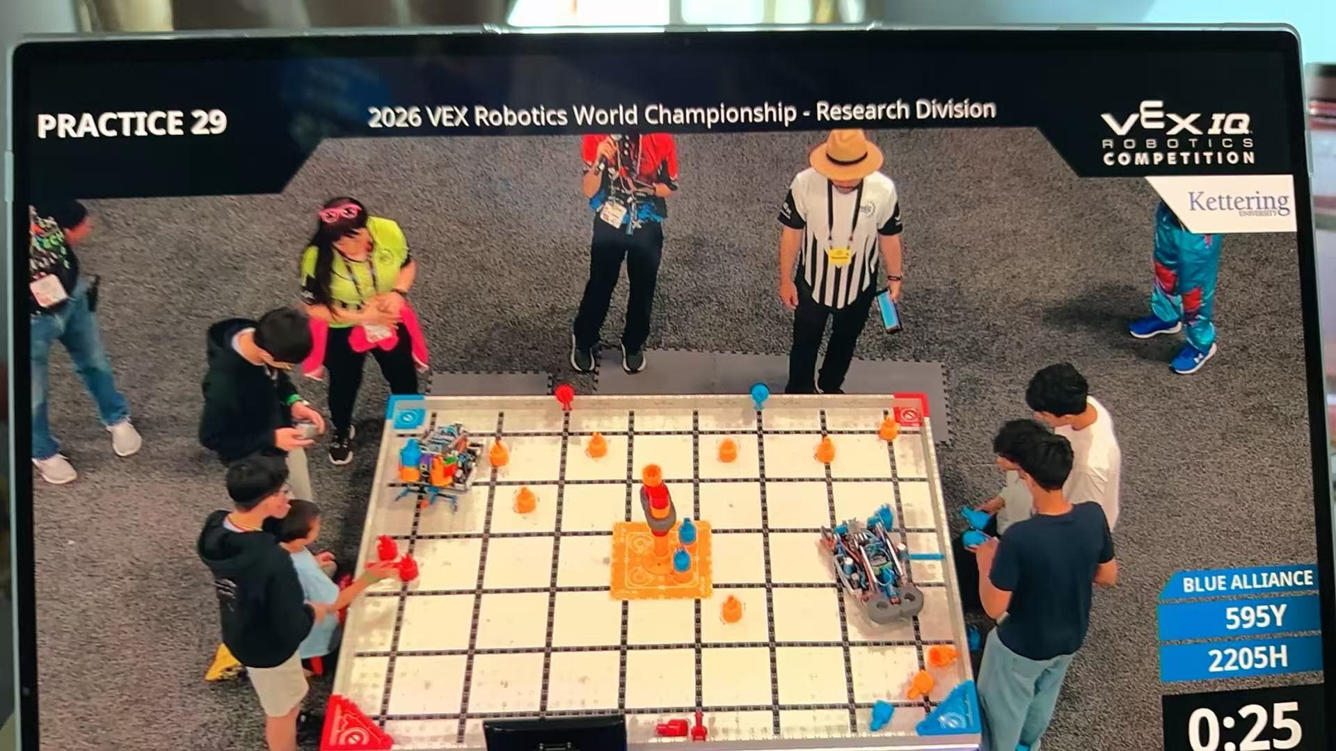 The screen above a World Championship field during a VEX IQ practice match, drivers standing around the field below.