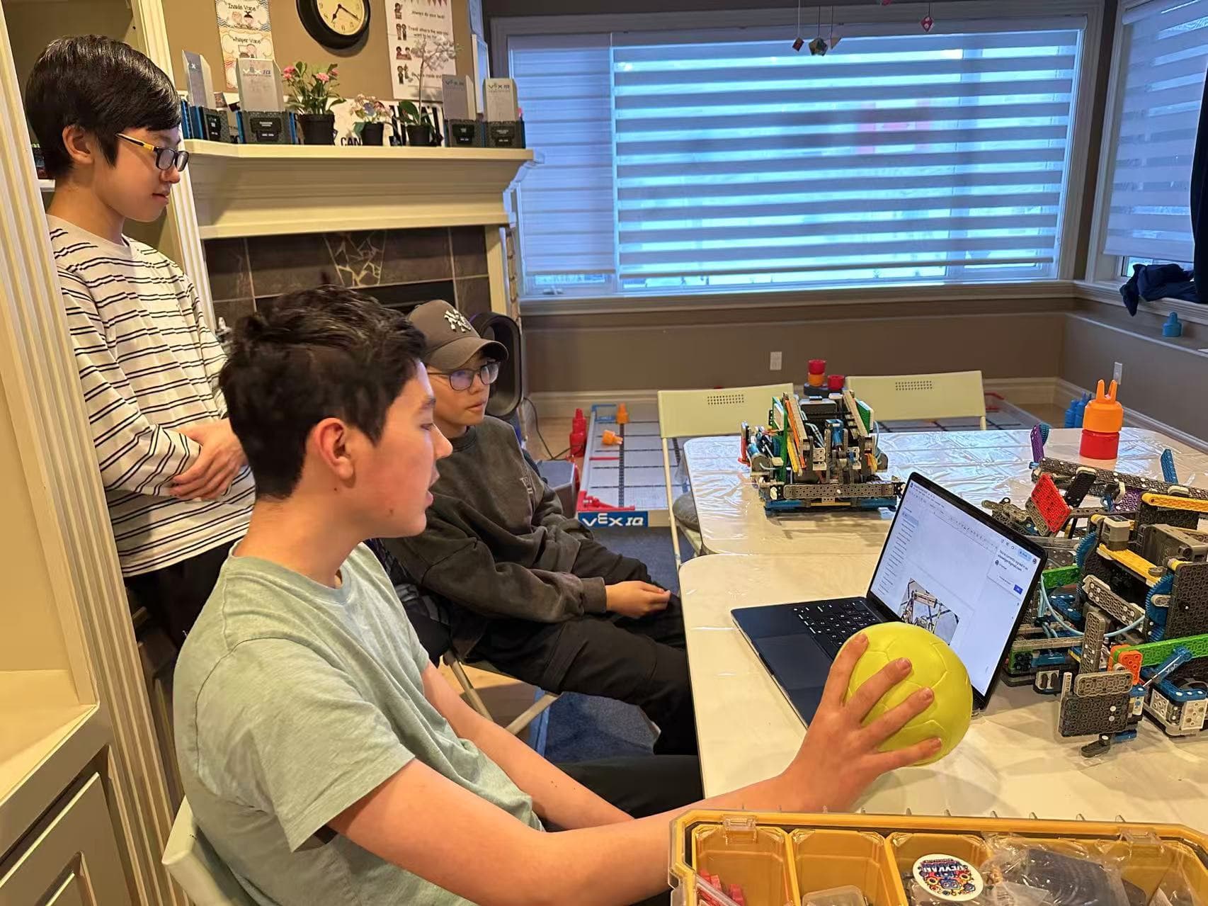 Three clubbers at a table with a VEX IQ practice field and a laptop in front of them, one of them holding a game ball.