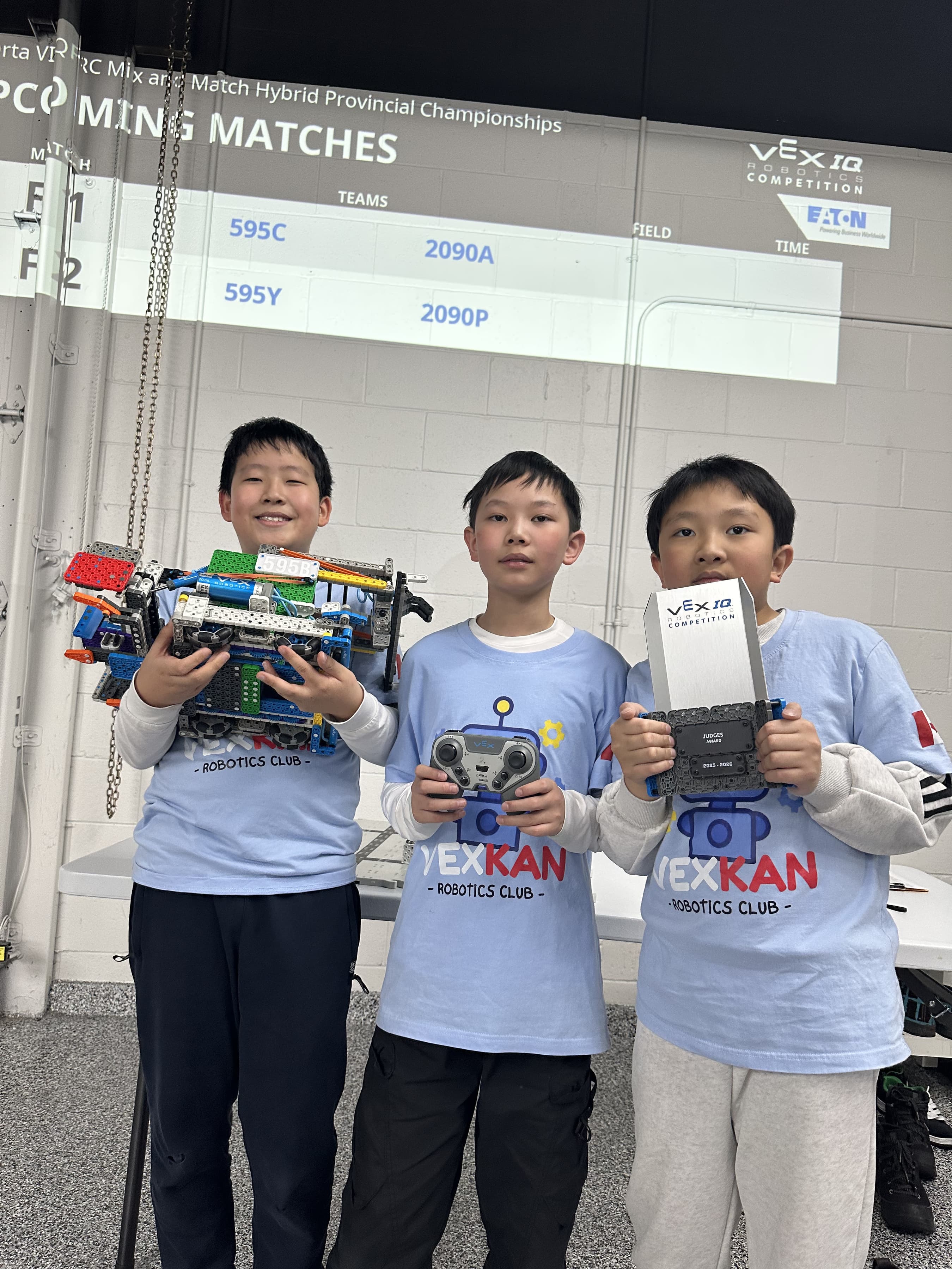 Three clubbers in club shirts holding their VEX IQ robot and a controller under the upcoming-matches screen at the provincial championships.