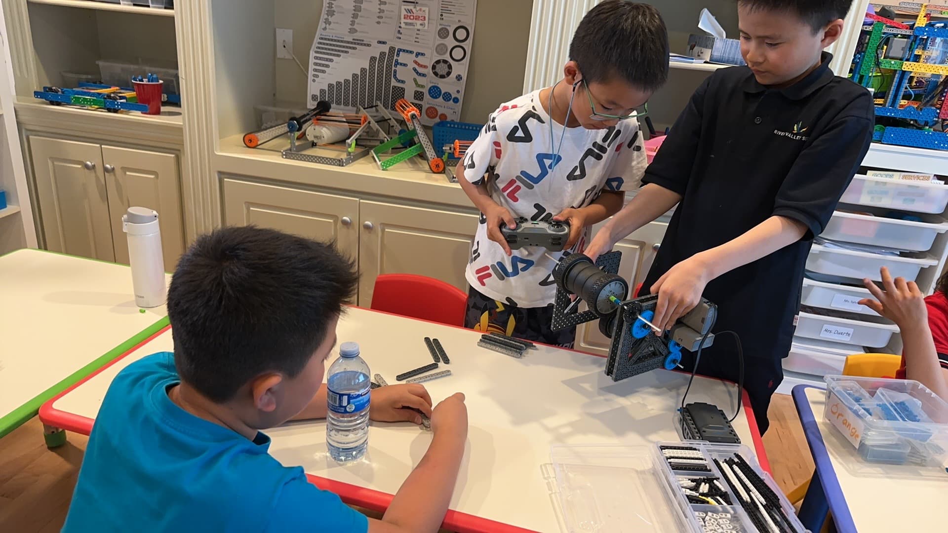 Three young clubbers around a classroom table, one driving a VEX IQ robot with a controller while another steadies it and a third sorts parts.