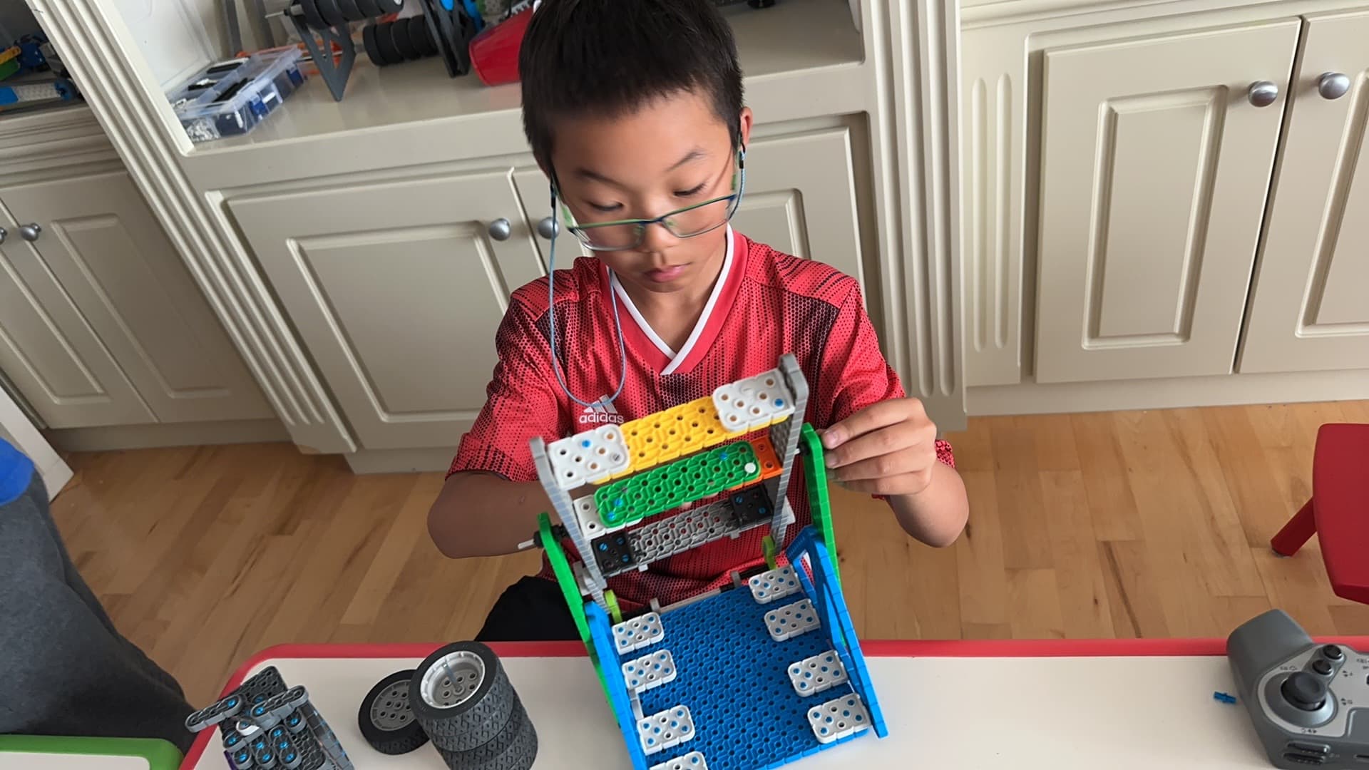 A young clubber holding up the VEX IQ robot he has just built, wheels and a controller on the table in front of him.