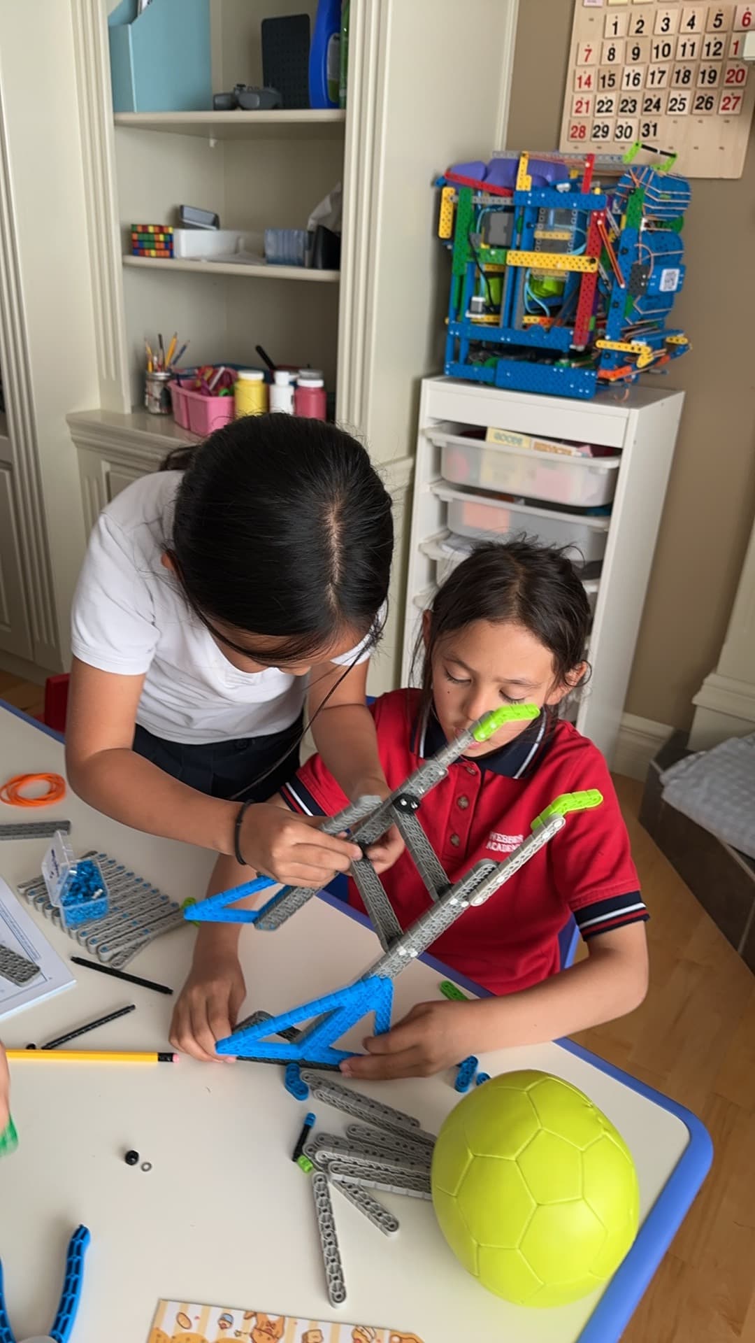 An older clubber leaning over a table to help a younger student fit a grey beam onto the blue VEX IQ frame she is holding, parts and a build guide spread out in front of them.