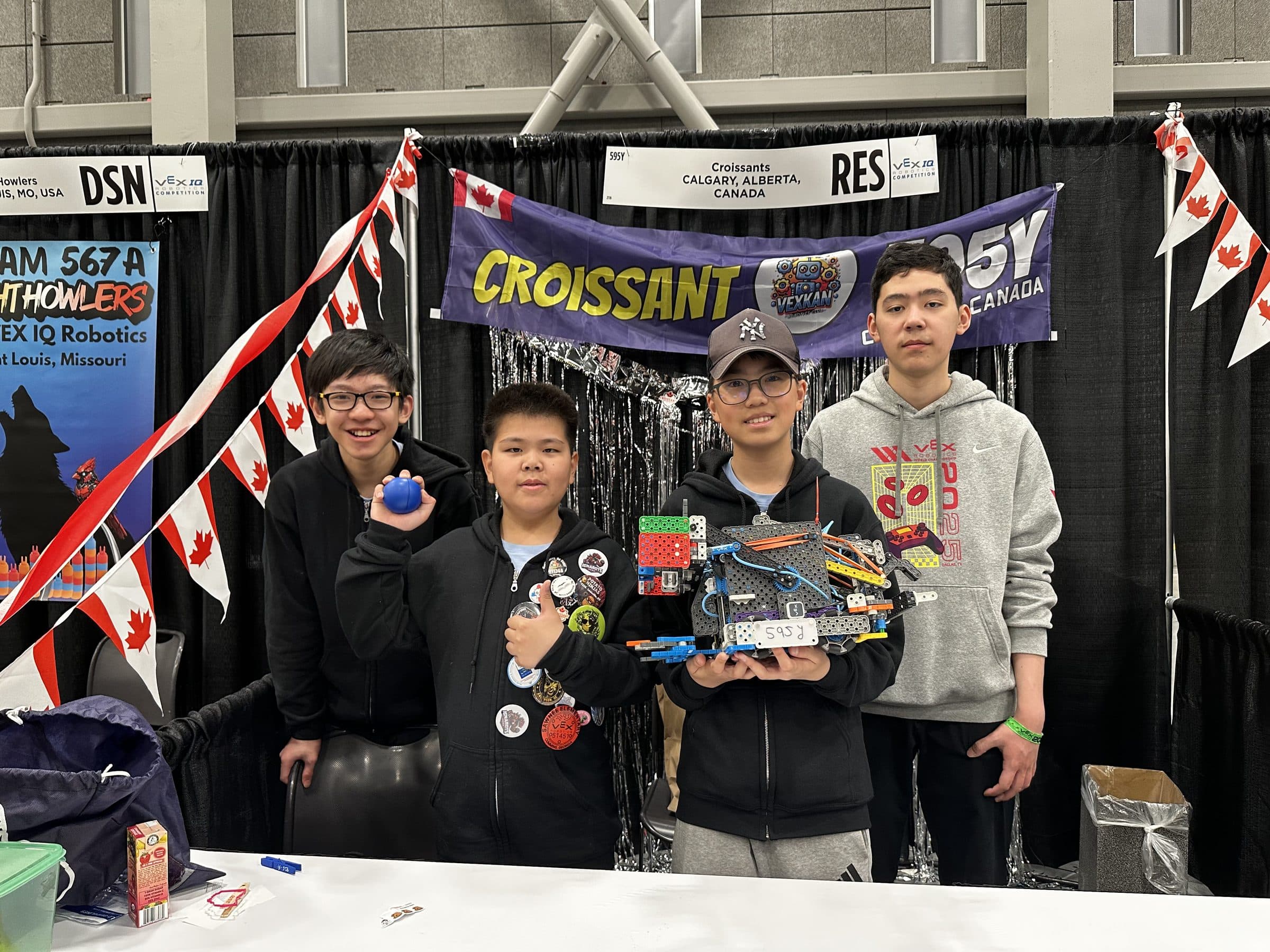 The four members of 595Y behind their pit table under the Croissants banner, one holding up the robot with 595Y written on its plate and another giving a thumbs up.