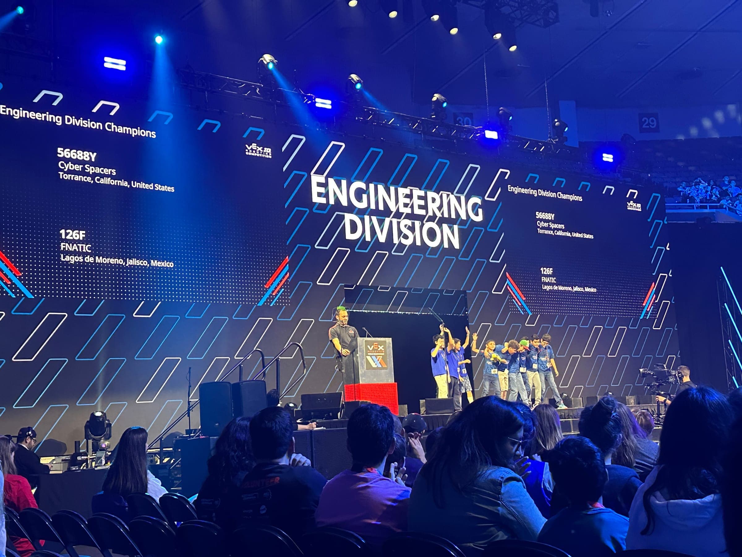 A division awards ceremony under way, the winning teams on stage and their team numbers listed on the screens either side.