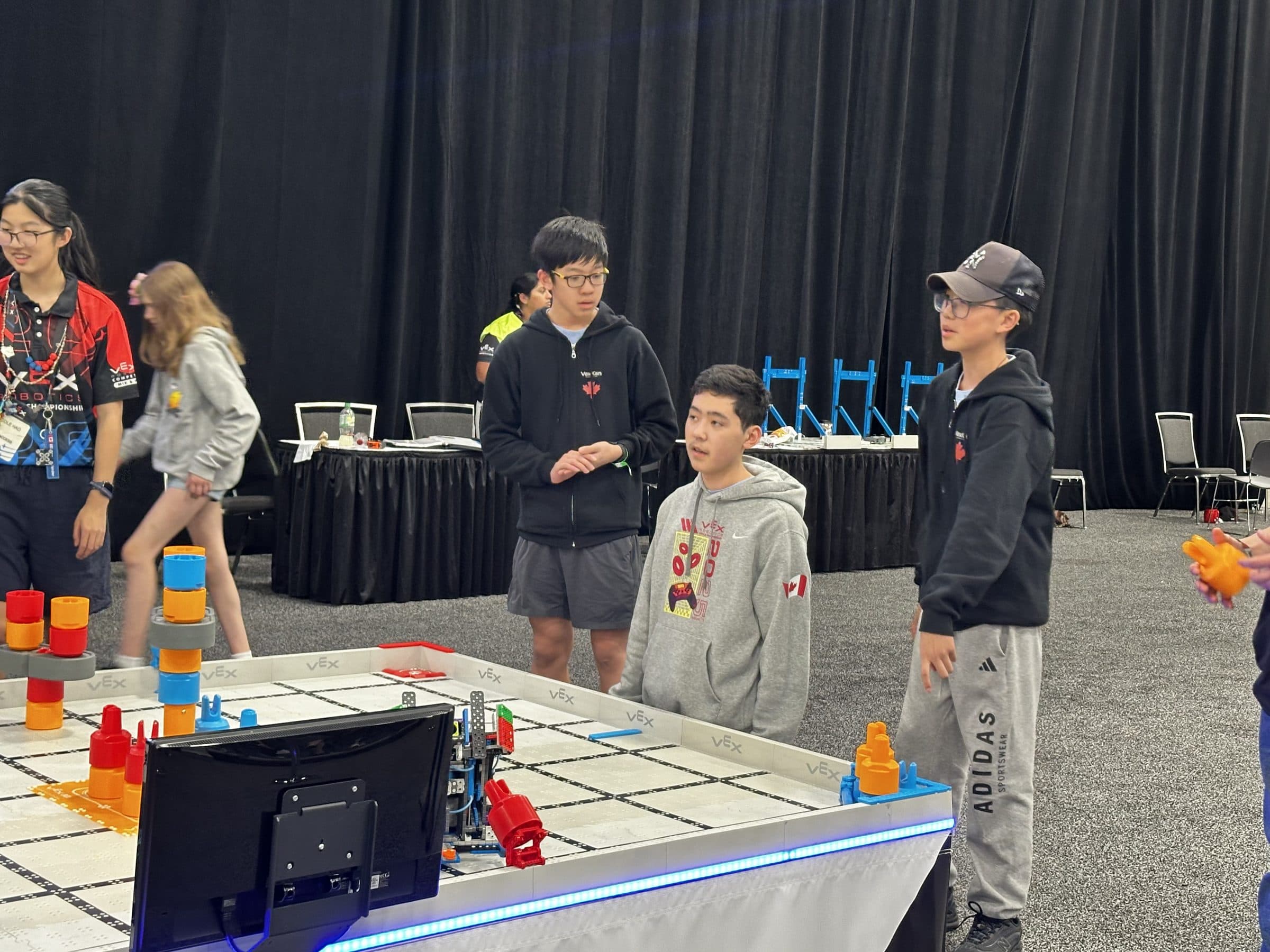 Three clubbers standing at the edge of a VEX IQ field between matches, talking through the next one with their robot already on the tiles.