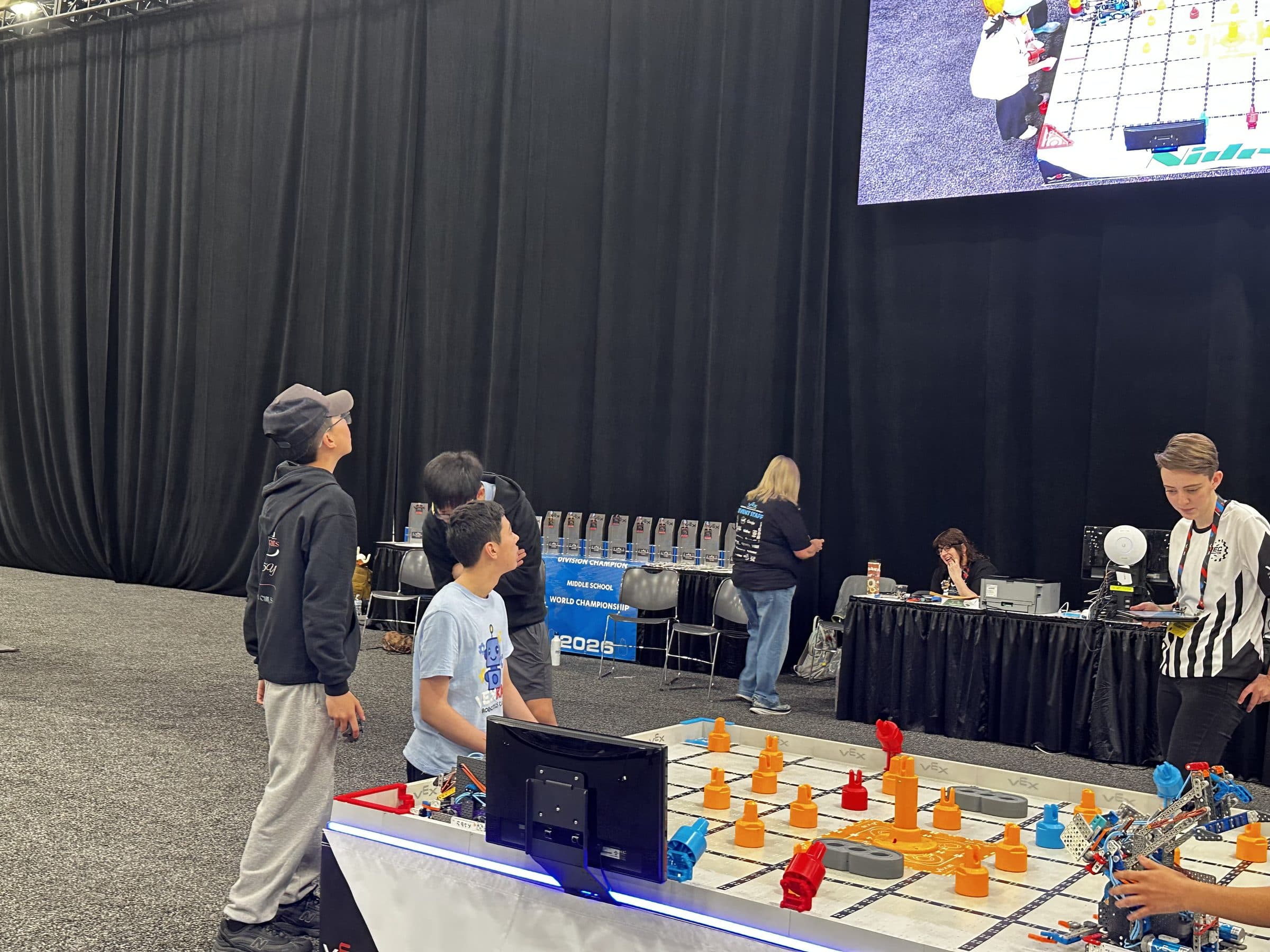 Clubbers waiting beside a VEX IQ field while a referee sets up, a row of Middle School World Championship division trophies laid out on the table behind them.