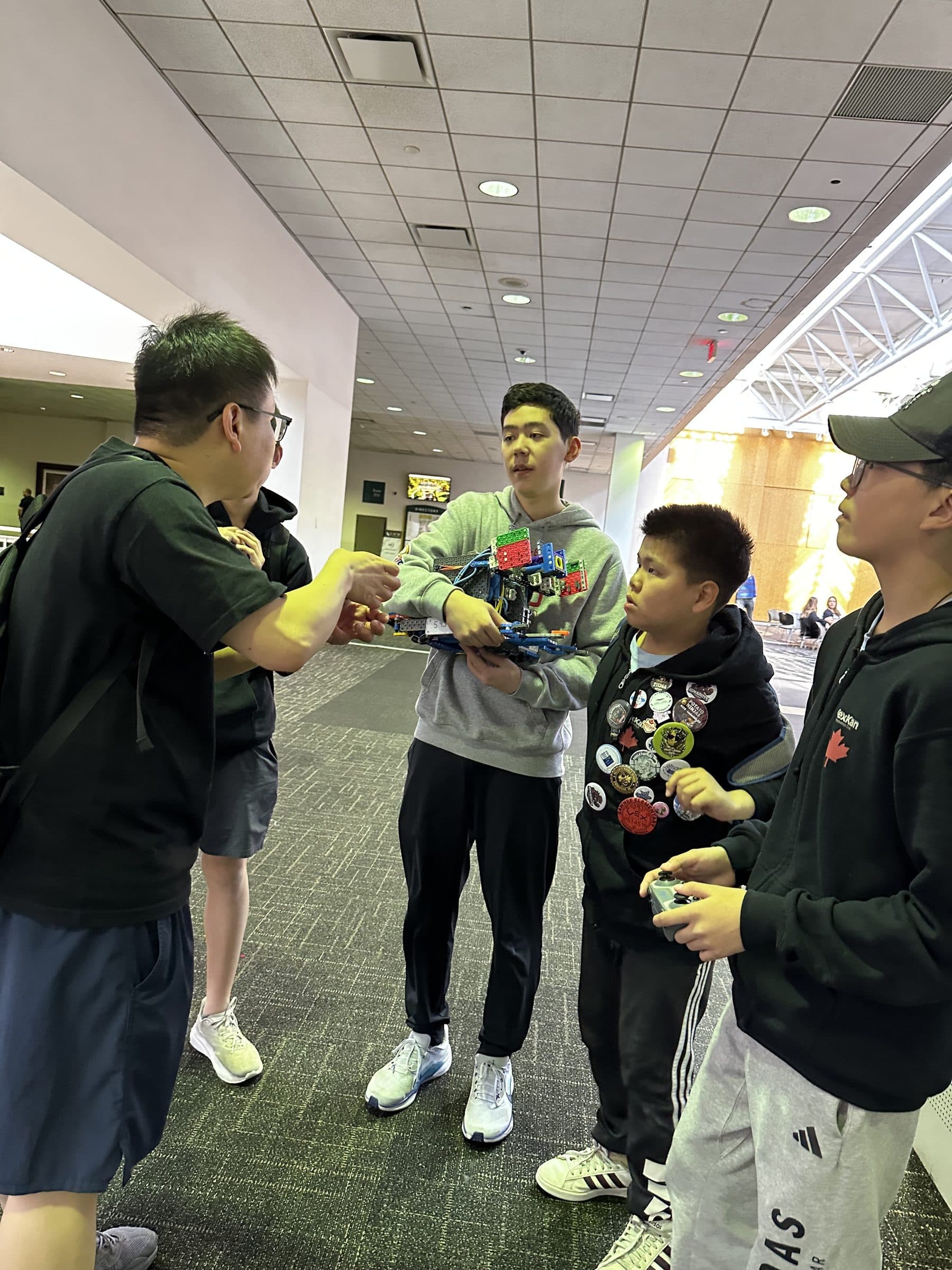 Clubbers gathered in the venue lobby, one holding a VEX IQ robot up to show the others, another wearing a lanyard covered in traded team pins.