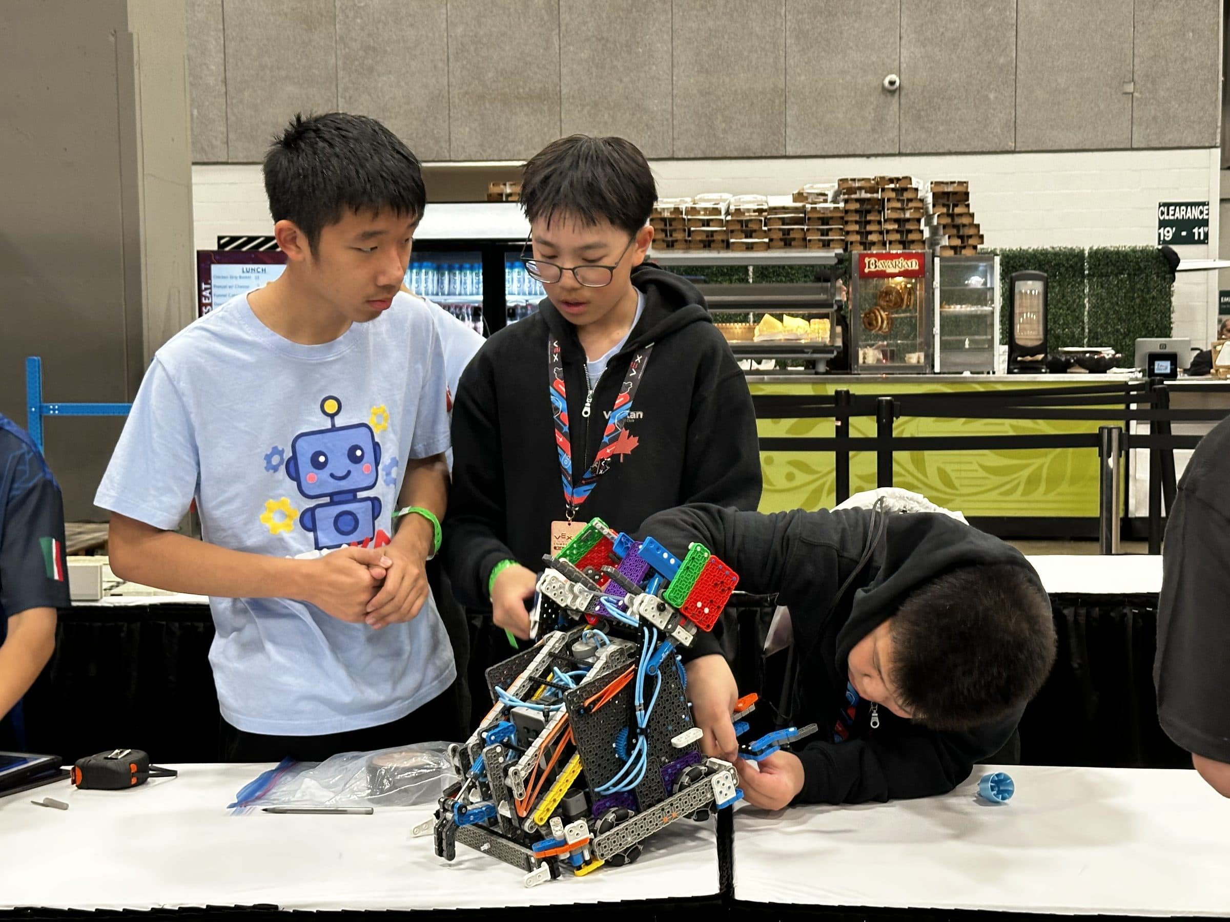Three clubbers rebuilding a robot on a table in the concourse, one leaning right over it to reach a part while the other two talk through what to do next.